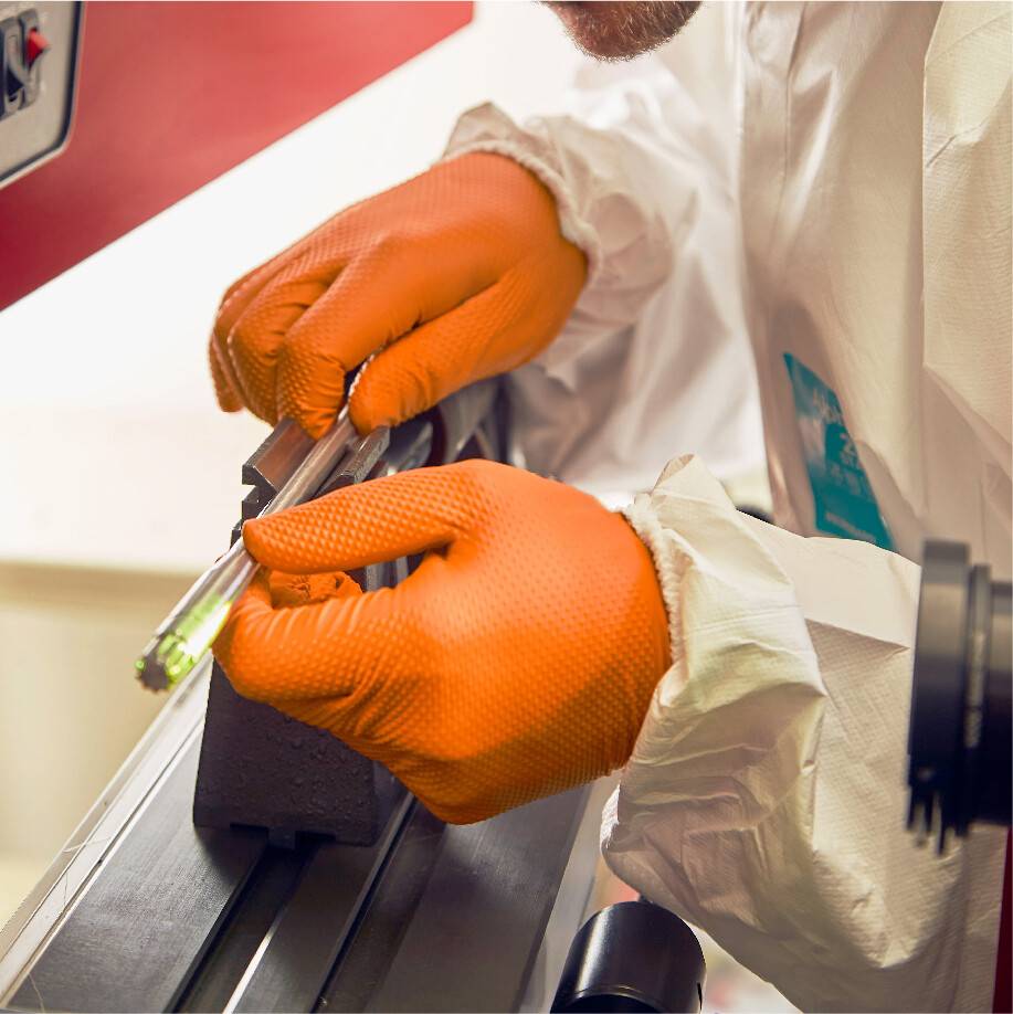 Osoba v ochranném oblečení a oranžových rukavicích opracovává kovovou součástku nástrojem v dílně.