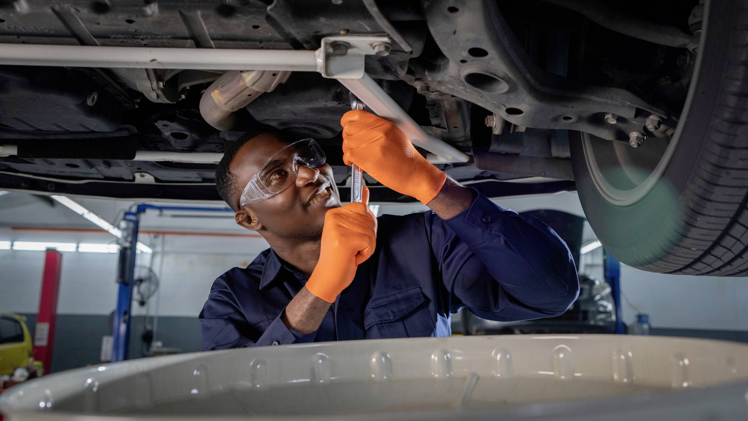 Mechanik v ochranných brýlích a oranžových rukavicích opravuje vozidlo zespodu v autoservisu.