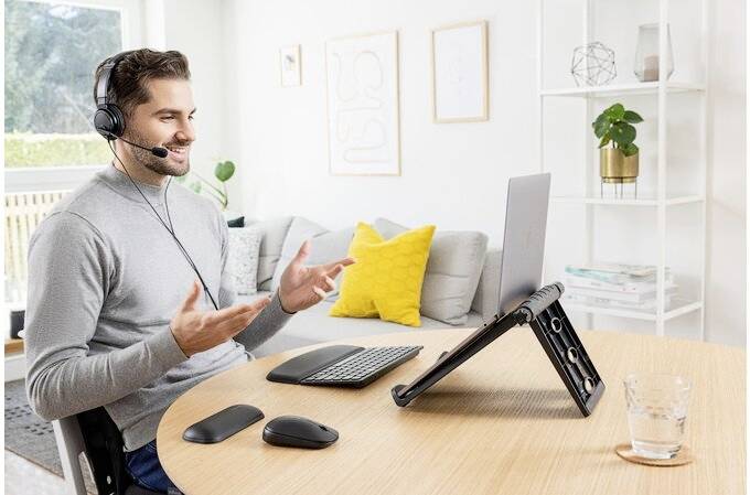 Muž se sluchátky sedí u stolu, mluví do laptopu a gestikuluje rukama. V pozadí jsou police a dekorace.