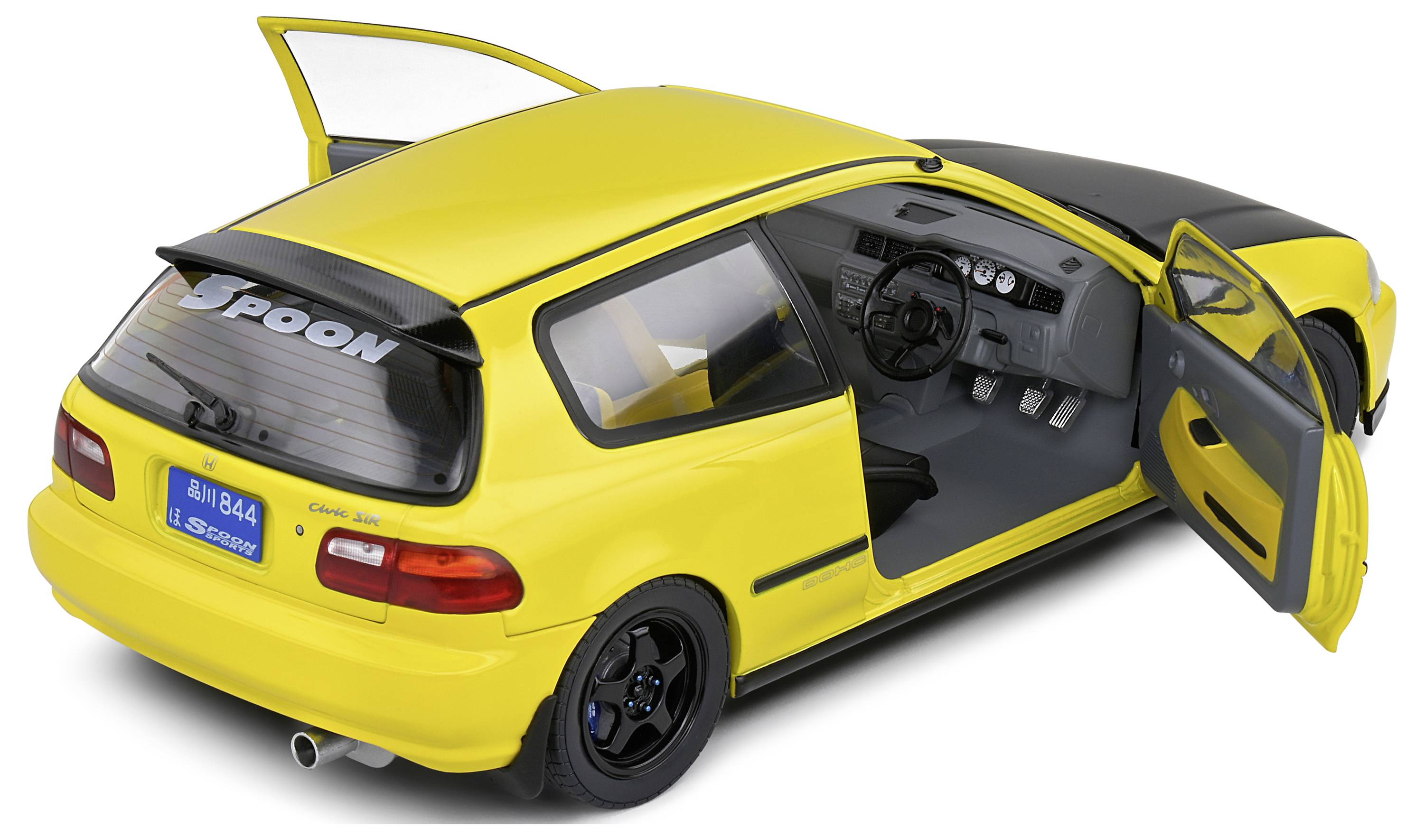 Žluté modelové auto v měřítku, s otevřenými dveřmi, ukazující volant a pedály, spoiler s nápisem 'Spoon', modrý držák poznávací značky.