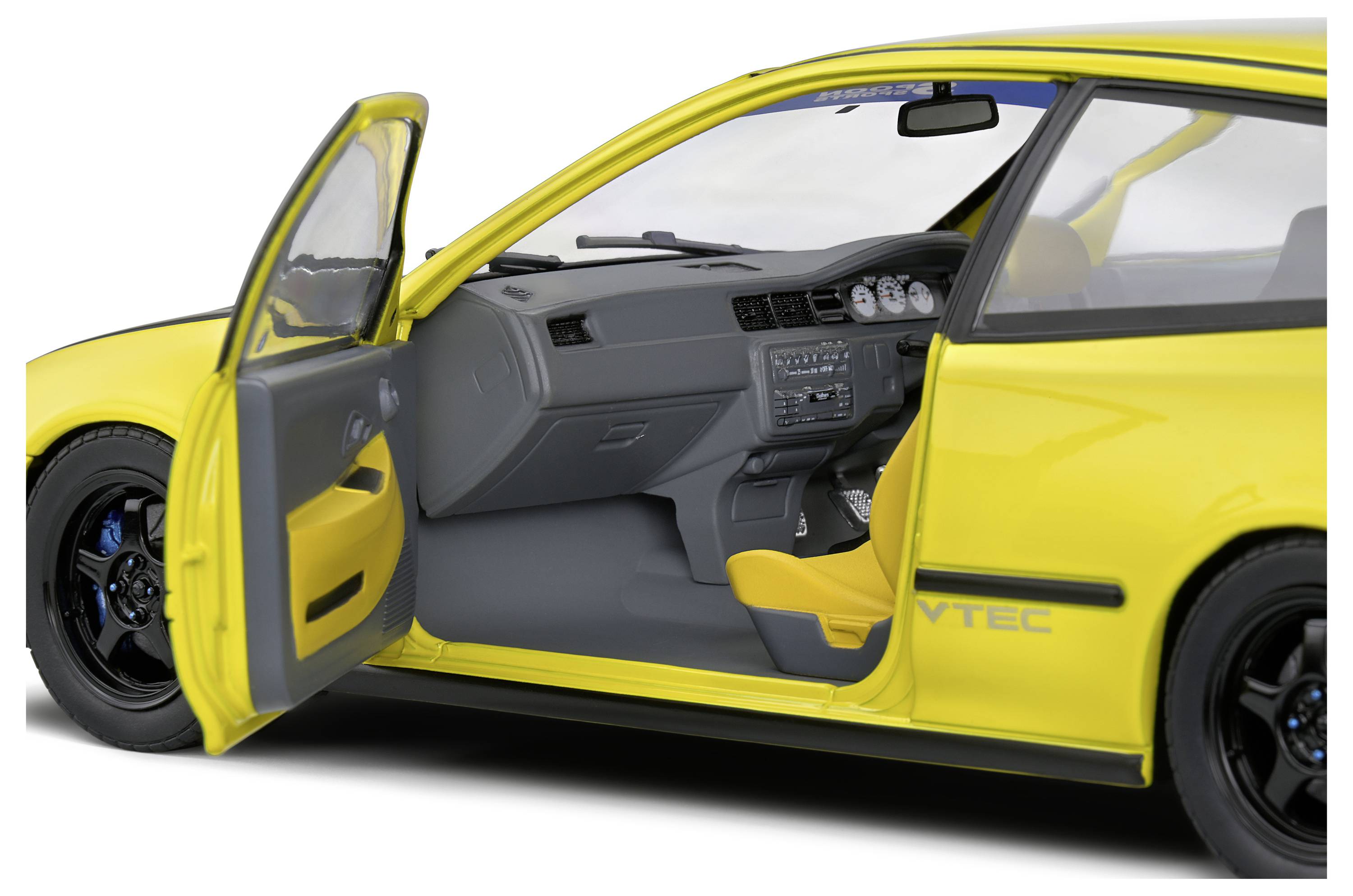 Žluté auto z boku s otevřenými řidičovými dveřmi, ukazuje šedý a žlutý interiér. Nápis 'VTEC' viditelný na karoserii.