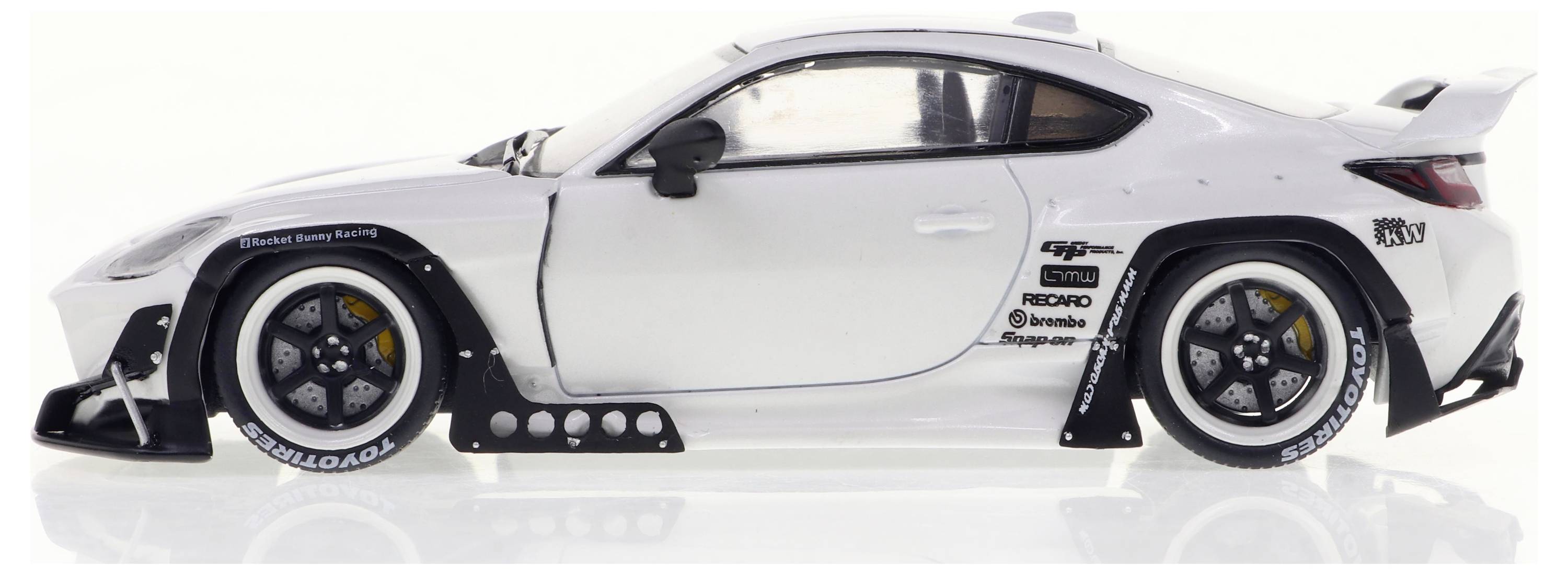 Bílý sportovní vůz z bočního pohledu s výraznými koly, spoilerem a závodními nálepkami. Automobil stojí před bílým pozadím.