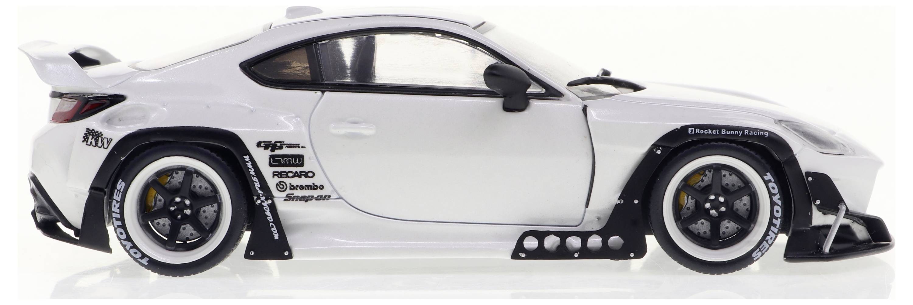 Bílé modelové auto se závodním designem, s viditelnými nápisy na pneumatikách a výraznými ráfky, pohled z boku.