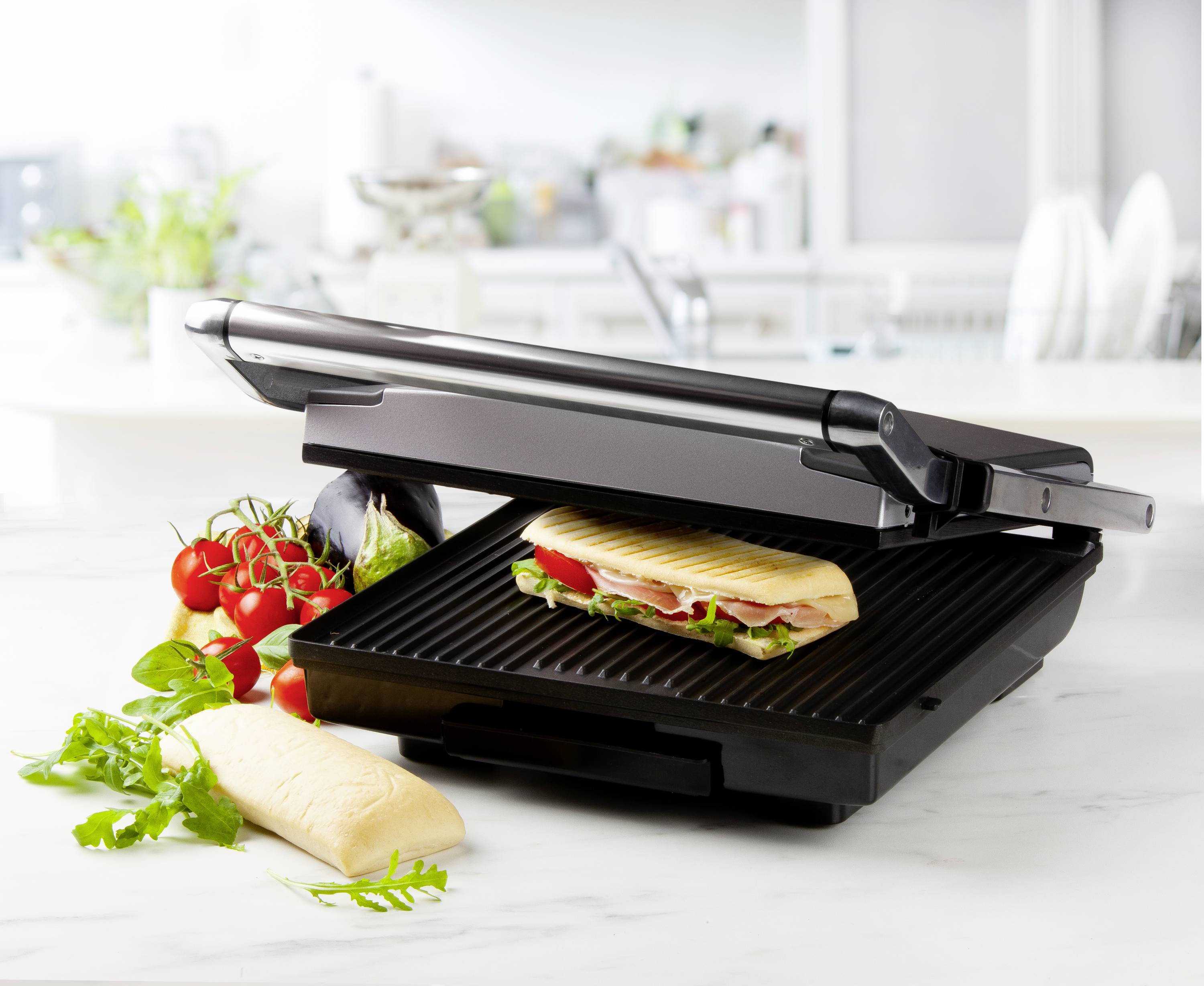 Otevřený kontaktní gril s panini a čerstvou zeleninou vedle něj na kuchyňské pracovní desce.