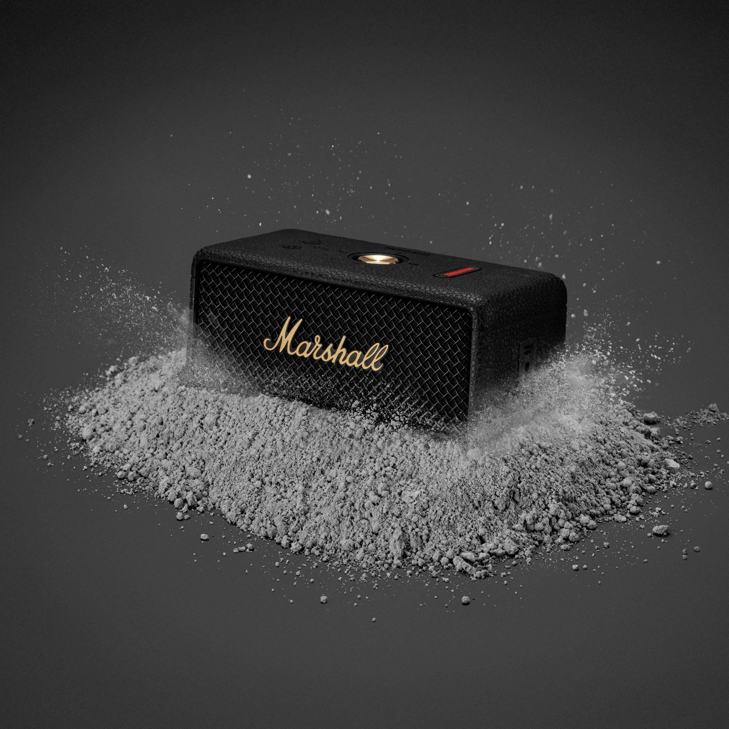 Černý bluetooth reproduktor ležící na hromadě popela, s nápisem "Marshall" zlatým písmem na přední straně.