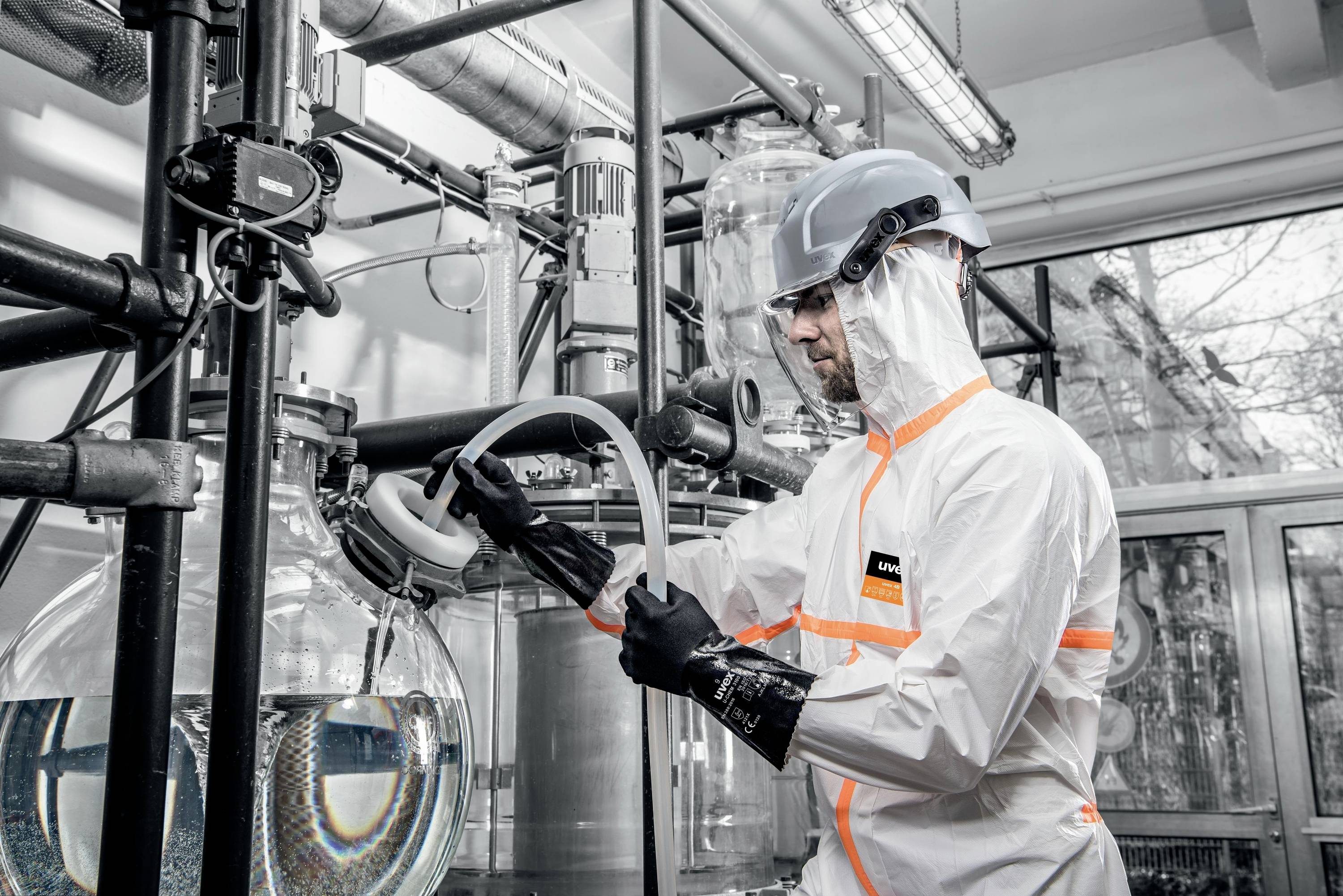 Laborant v ochranném oblečení a helmě pracuje v laboratoři u skleněného zařízení s velkými baňkami obsahujícími kapaliny.