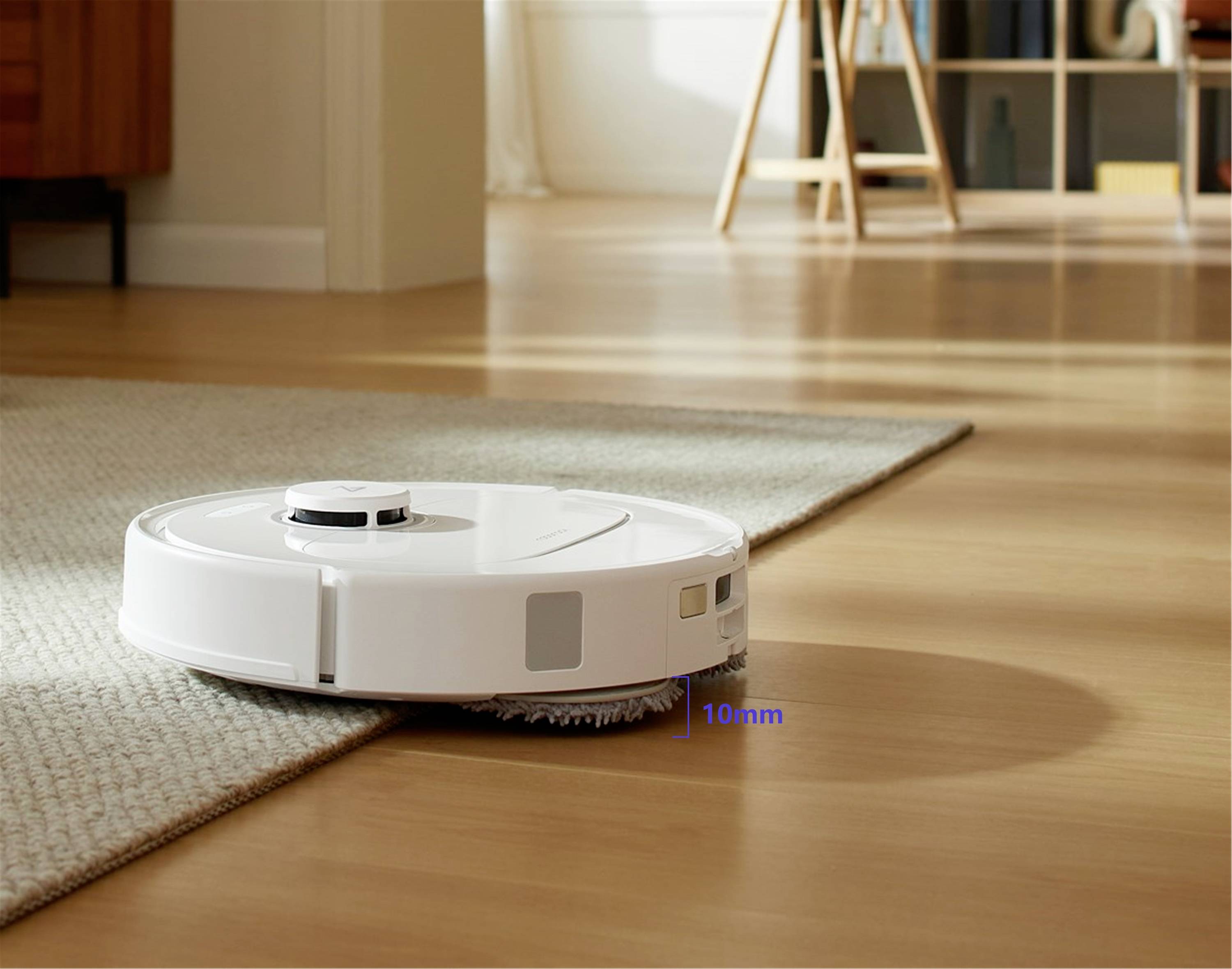 Bílý robotický vysavač přejíždí z dřevěné podlahy na koberec. V pozadí je vidět útulný interiér obývacího pokoje.
