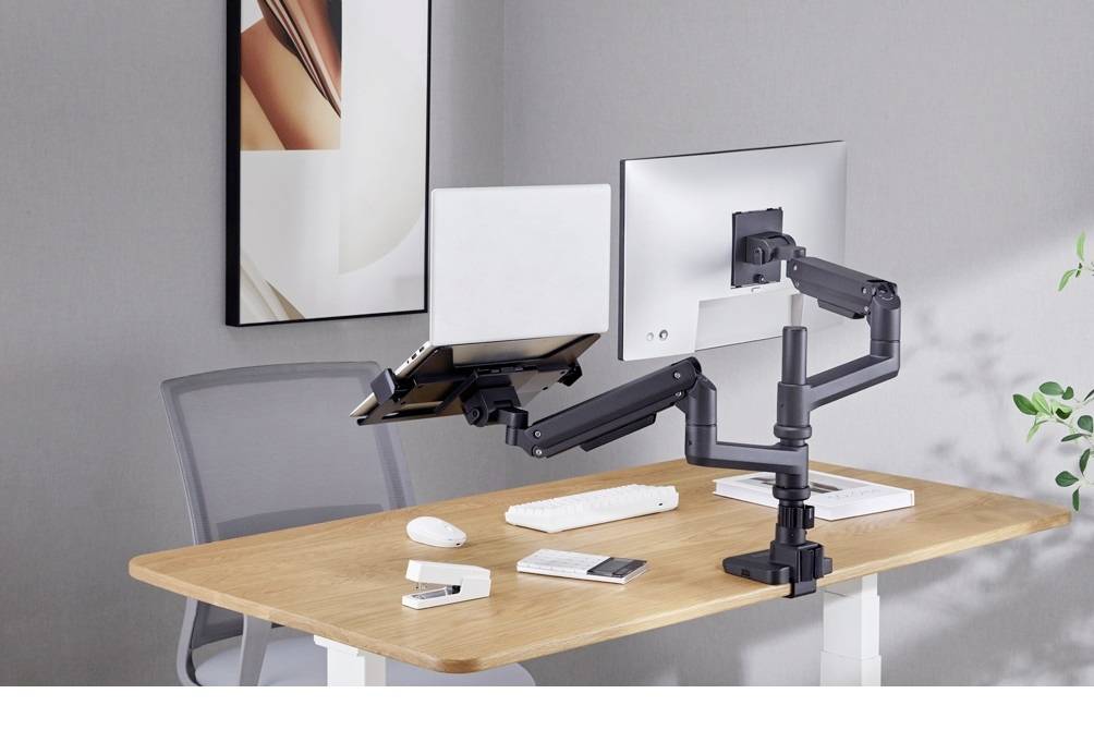 Psací stůl s dvěma monitorovými rameny, která drží laptop a monitor. V pozadí šedá židle a abstraktní umělecký obraz.