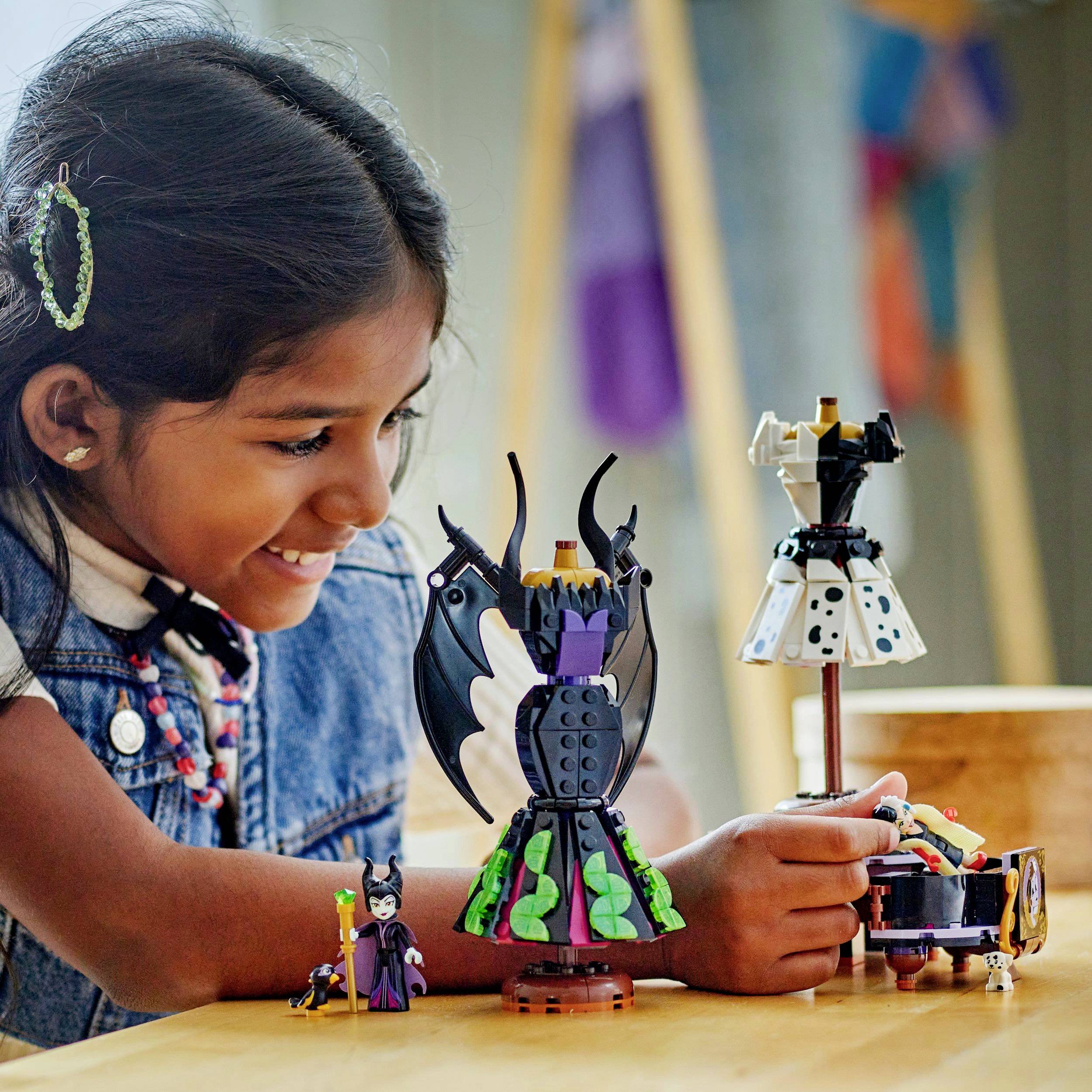 Dívka se usmívá a hraje si s barevnými malými robotickými figurkami na stole. Roboti mají propracované a umělecké designové prvky.