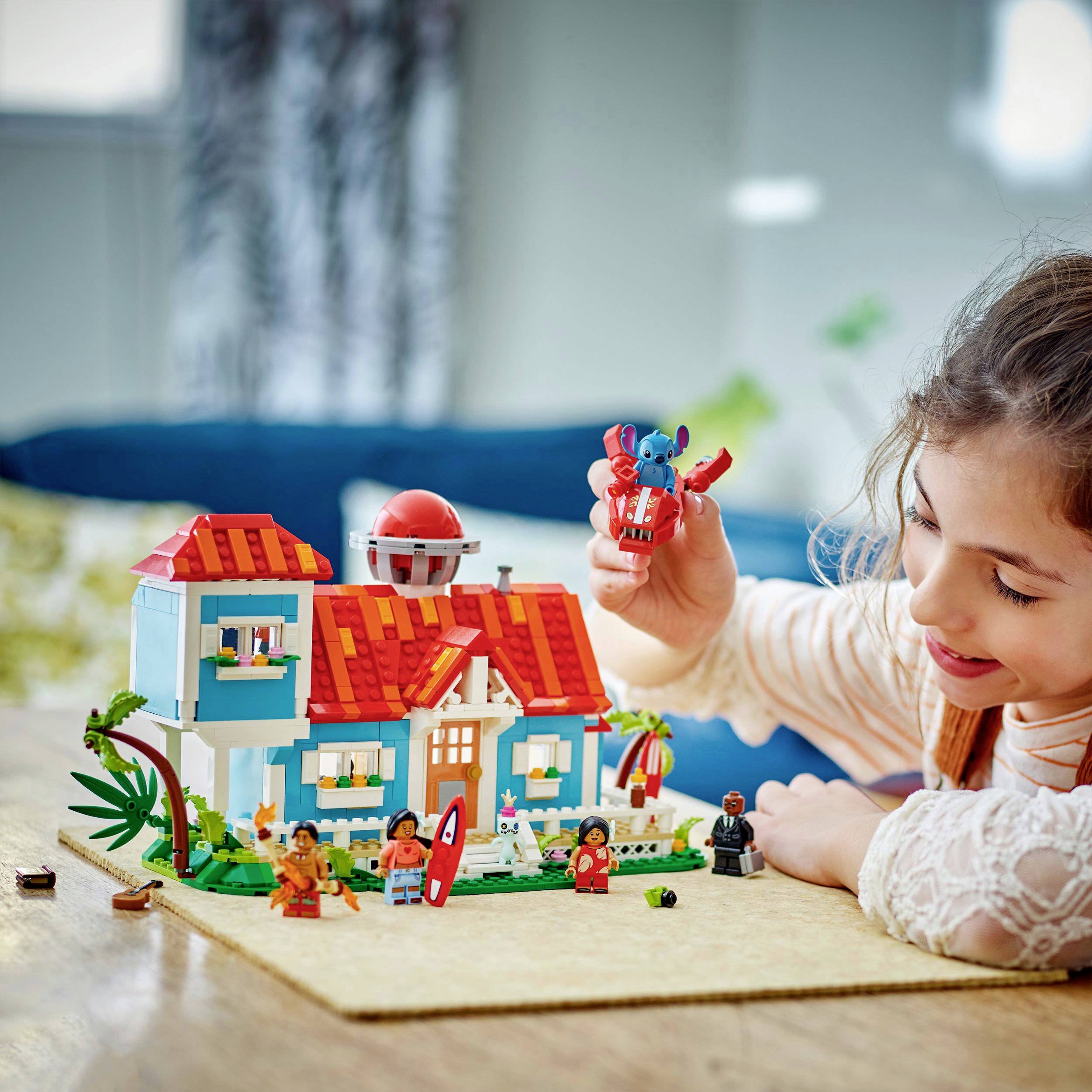 Dítě si hraje s Legem, drží figurku nad pestrobarevným legodomem. Figurky stojí kolem domu, který je postaven na stole.