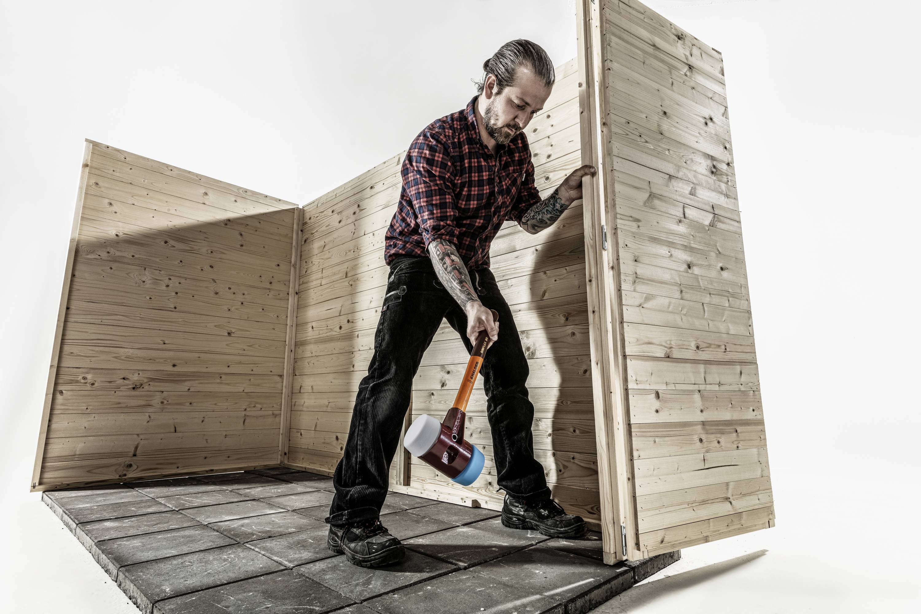 Muž s vousy a tetováním staví dřevěnou stěnu na dlážděné podlaze; řemeslnická práce s nářadím.