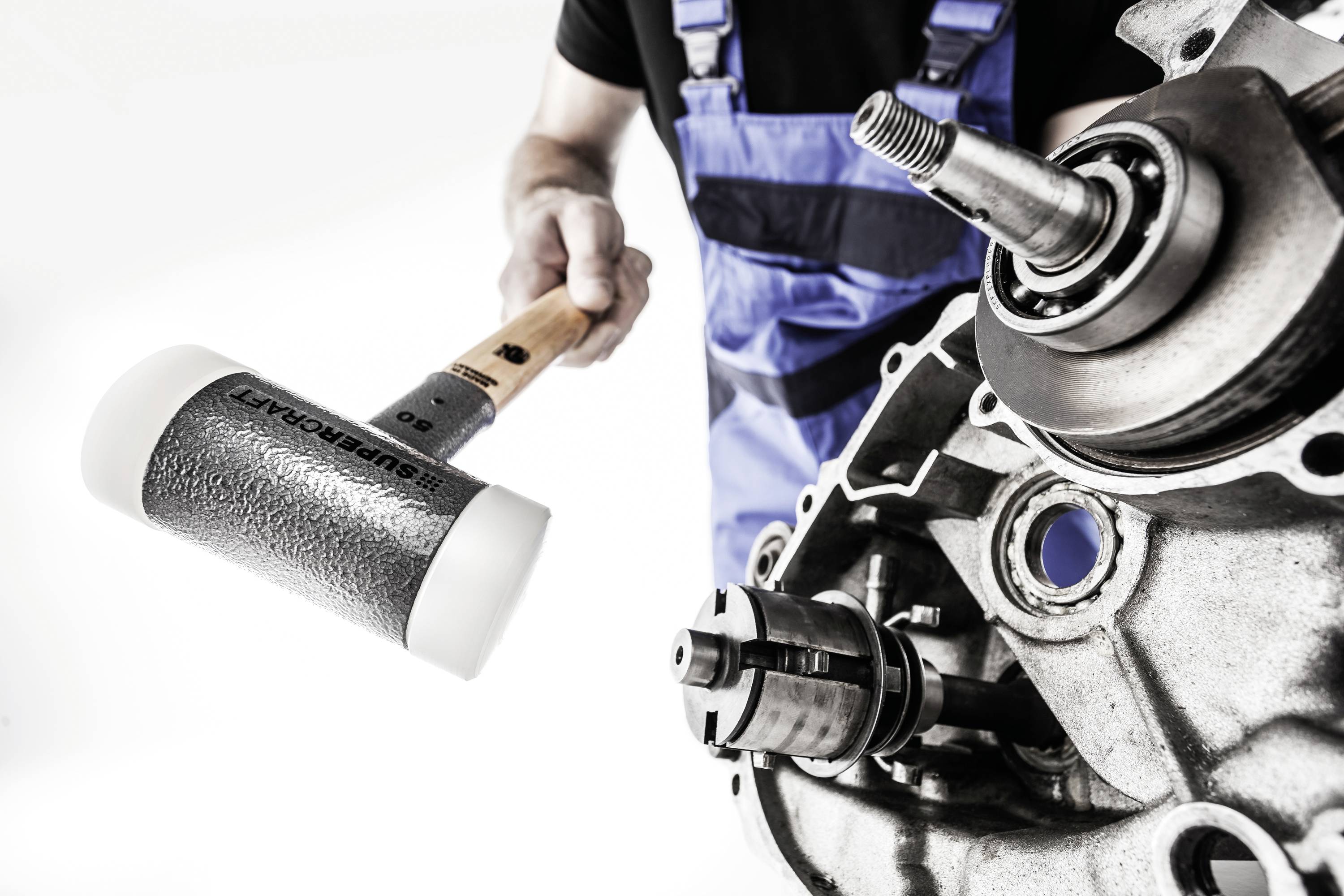 Mechanik v modré pracovní kombinéze drží gumovou palici vedle části motoru, připravený na opravárenské práce.