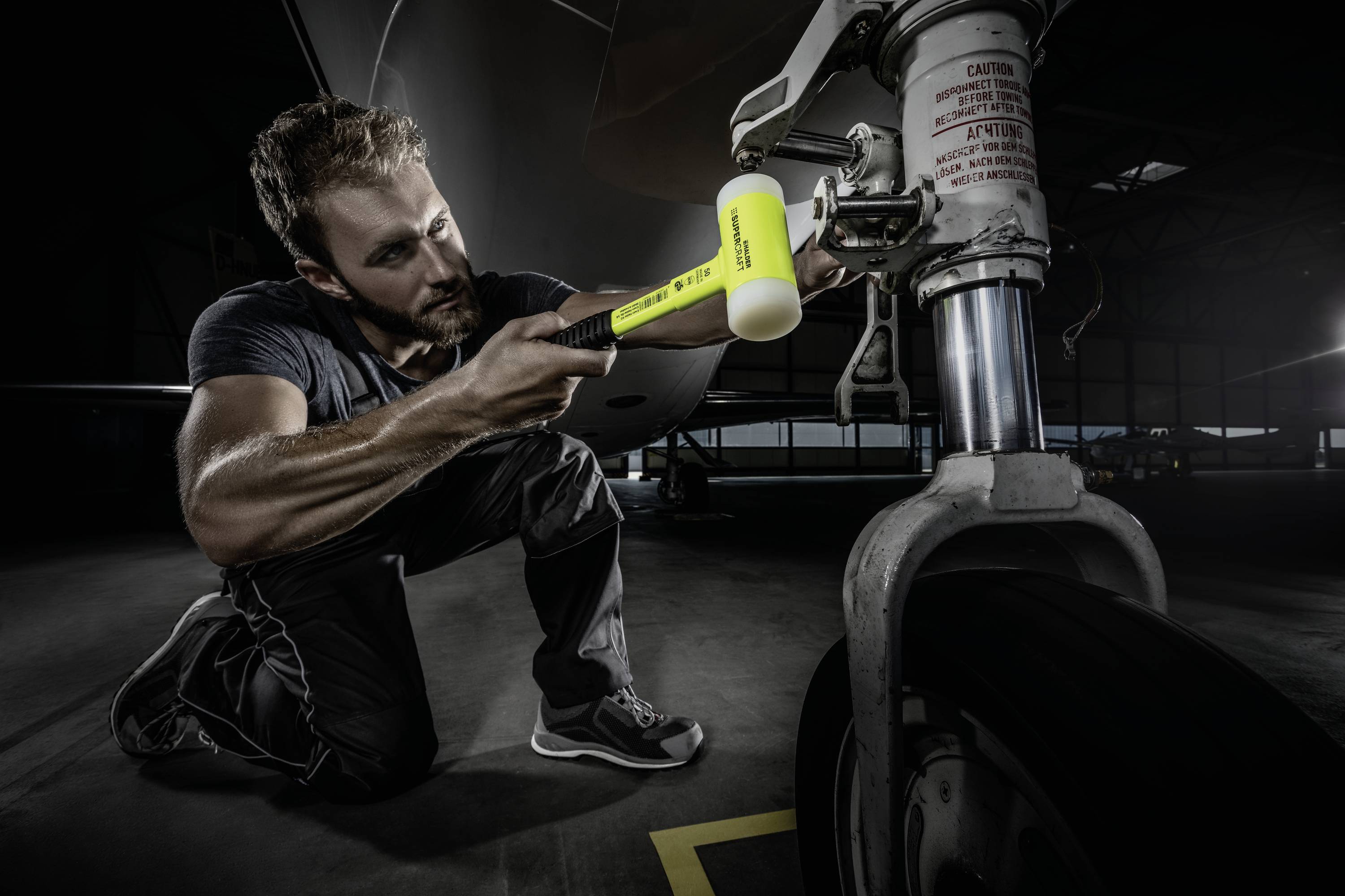 Mechanik klečí a používá žlutý kladívko ke kontrole podvozku letadla. Soustředěně se věnuje své práci.