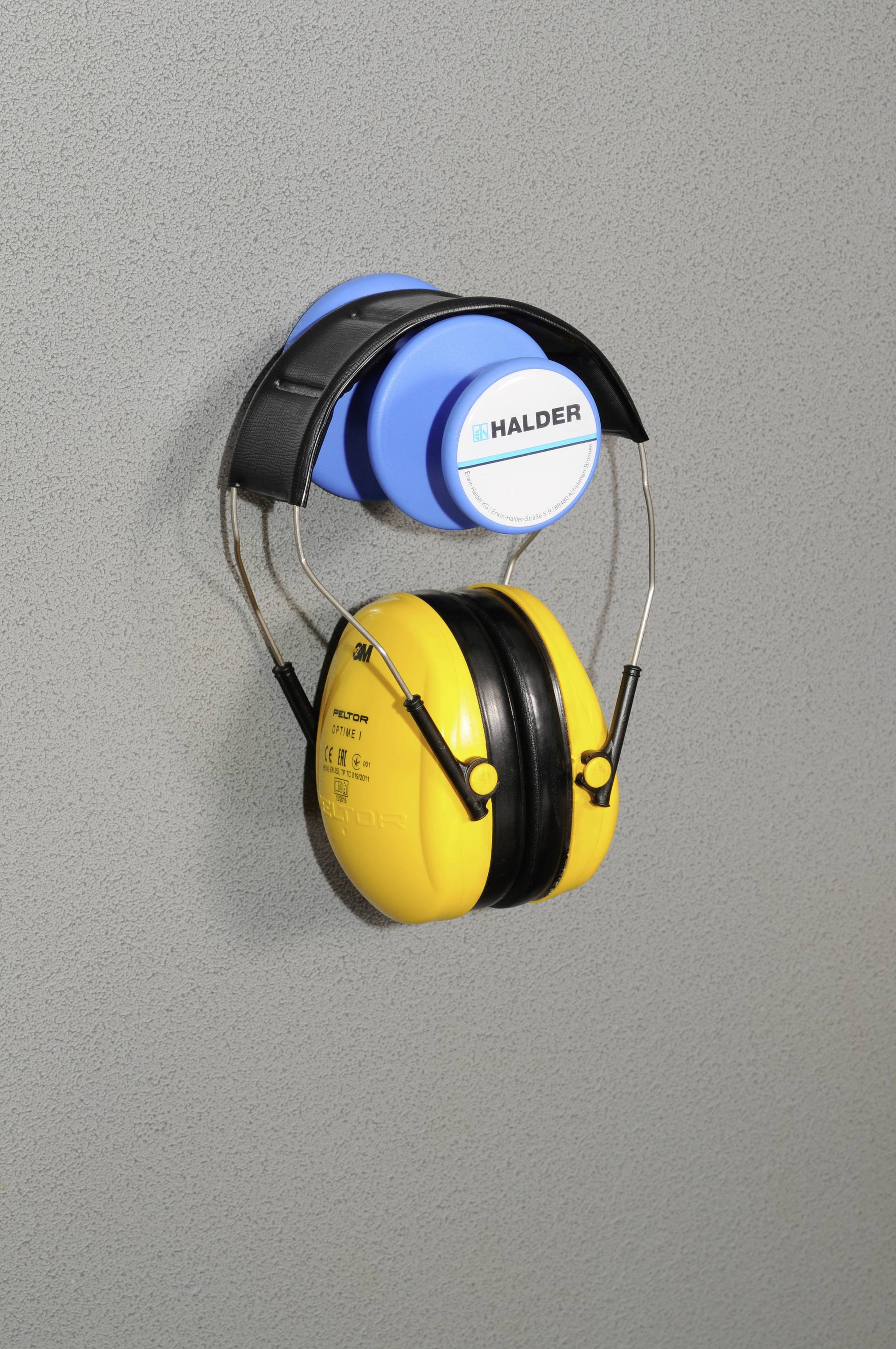 Žluté chrániče sluchu visí na šedém nástěnném držáku značky 'Halder'. Jsou navrženy pro ochranu před hlukem a bezpečnost práce.