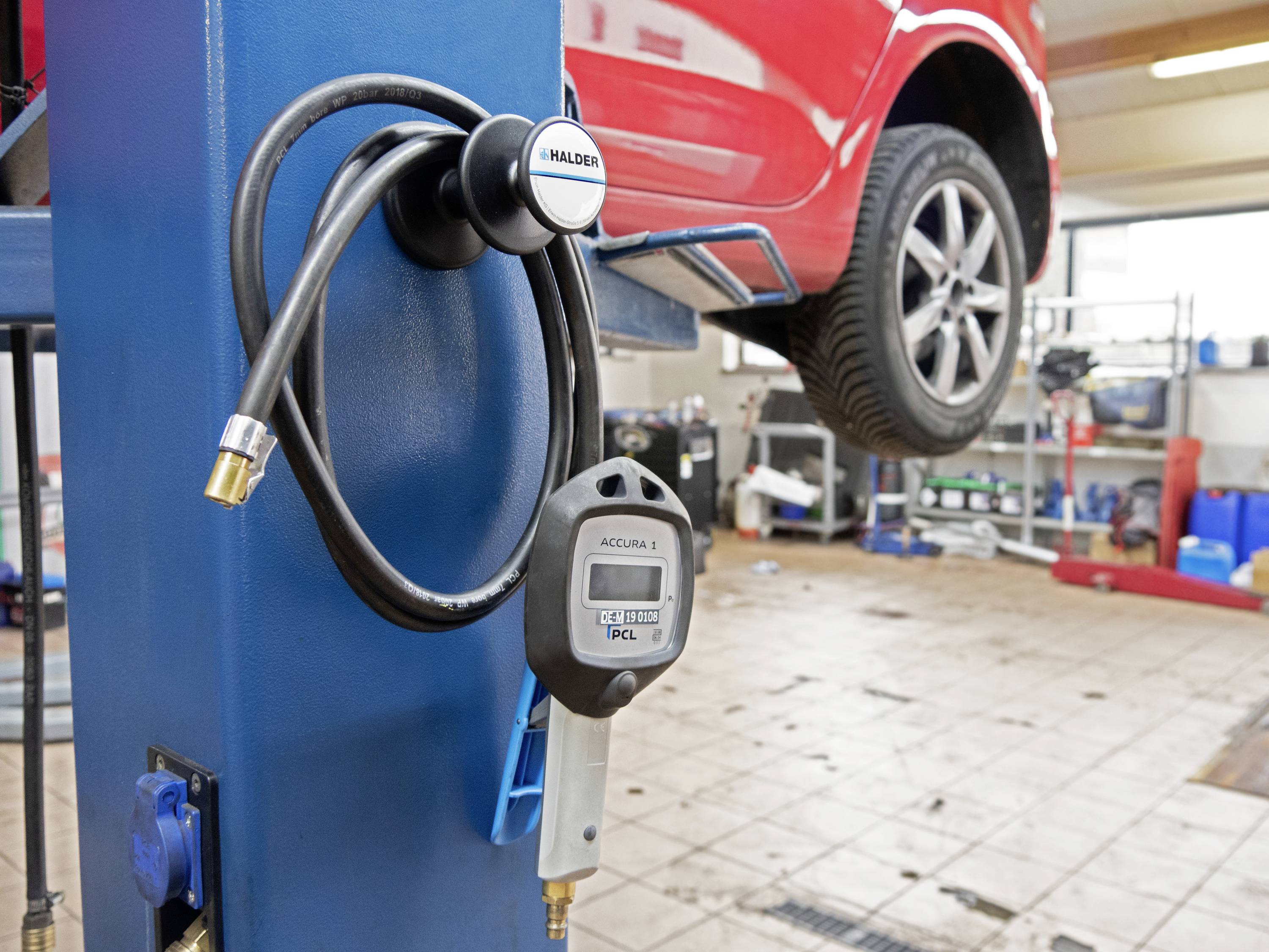 Červené auto je zvednuté na zvedáku v autoservisu. V popředí visí tlakoměr a hadice na modrém sloupu.
