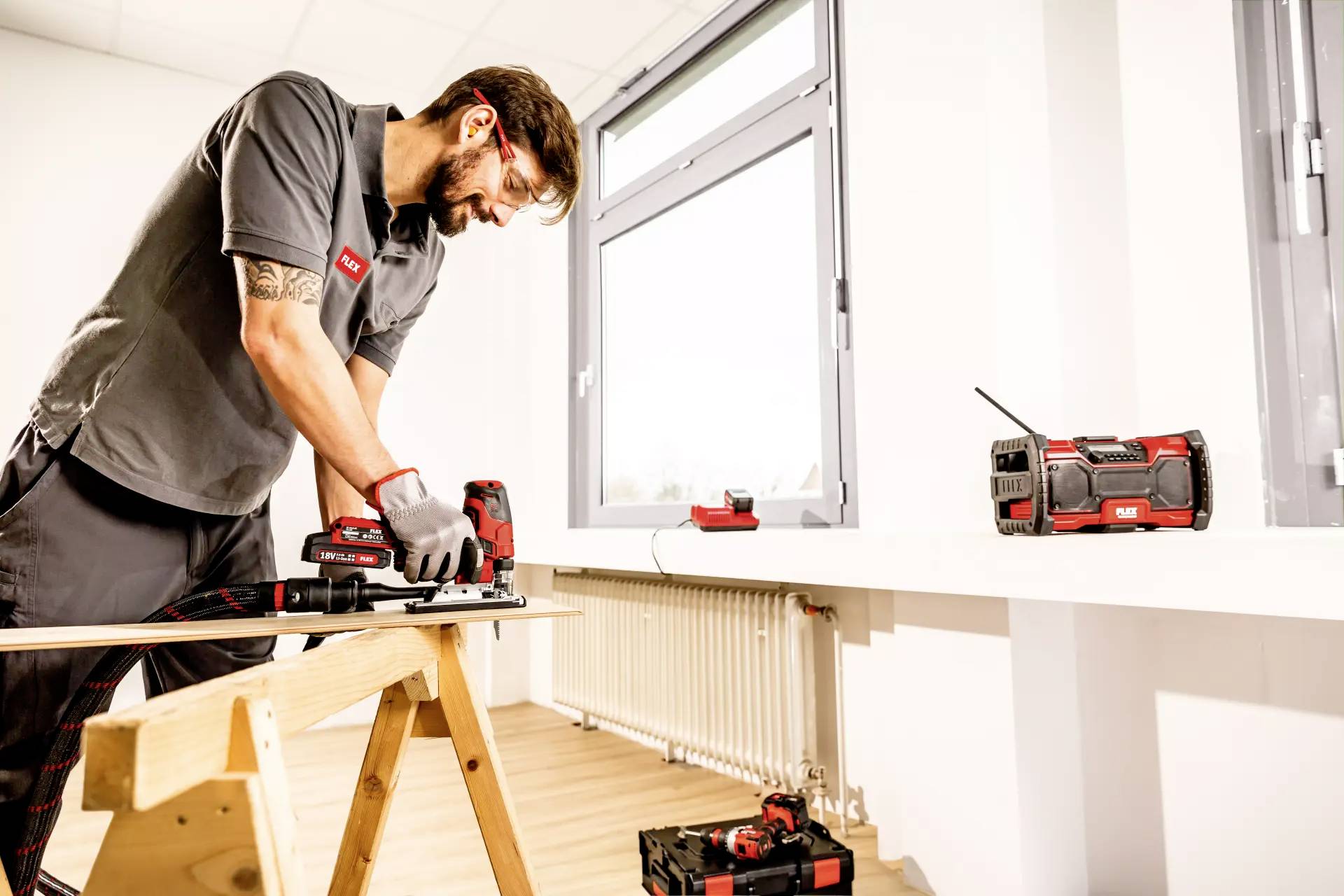 Muž v pracovním oblečení používá elektrickou pilu na pracovním stole v dobře osvětlené místnosti. V okolí jsou viditelné nástroje a rádio.