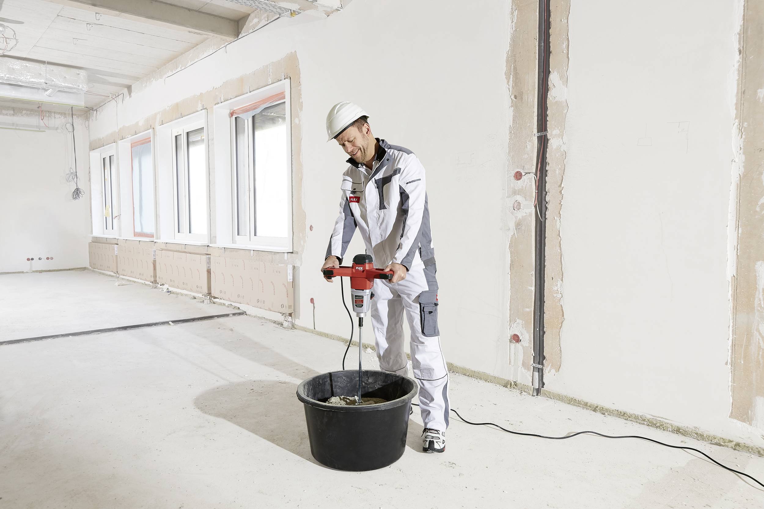 Dělník v ochranném oblečení míchá beton elektrickým míchadlem v prázdné místnosti s částečně odhaleným zdivem.