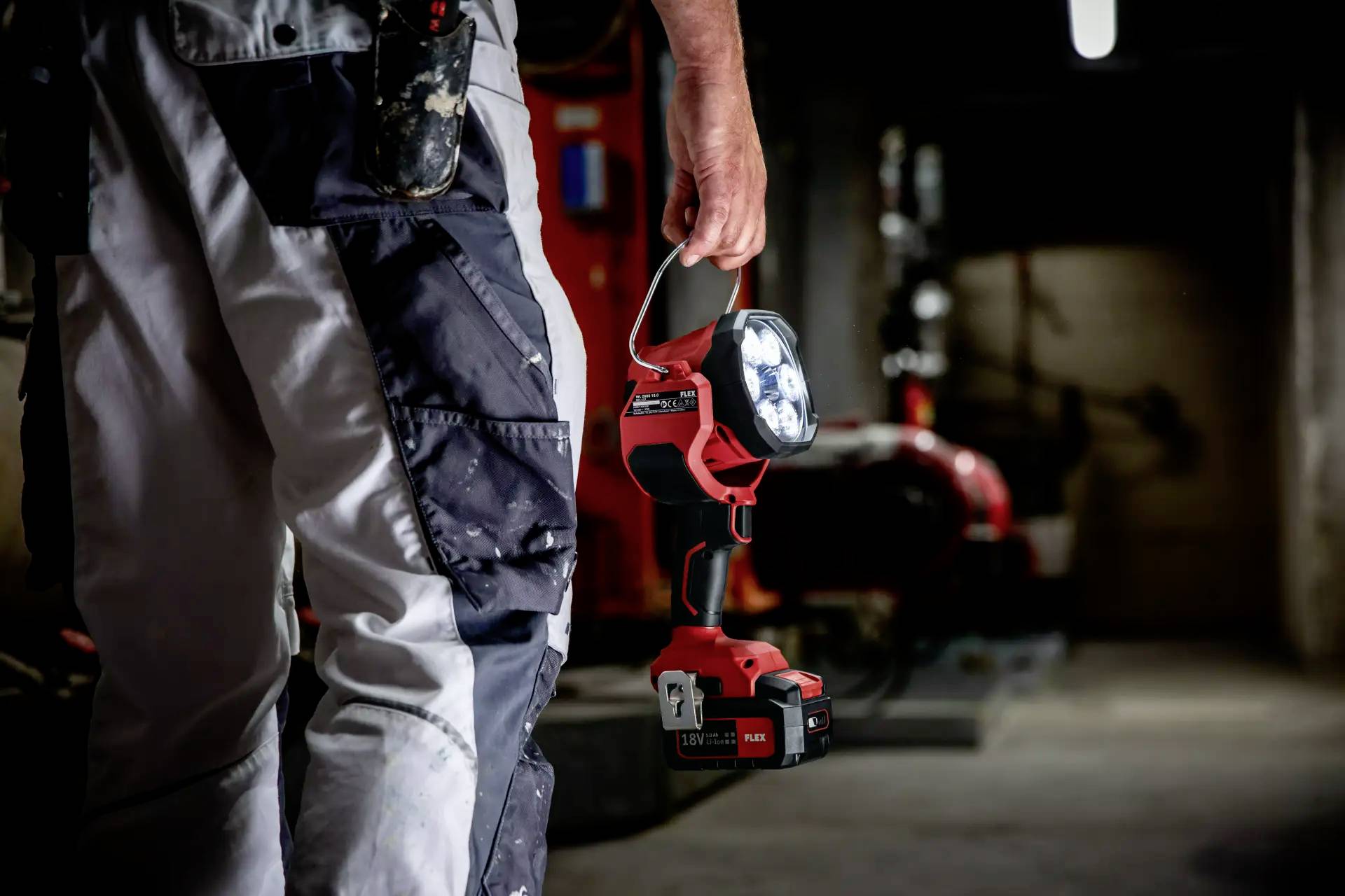 Dělník v pracovním oblečení drží přenosnou LED lampu v ponurém, industriálním sklepě. V pozadí jsou viditelné velké stroje.