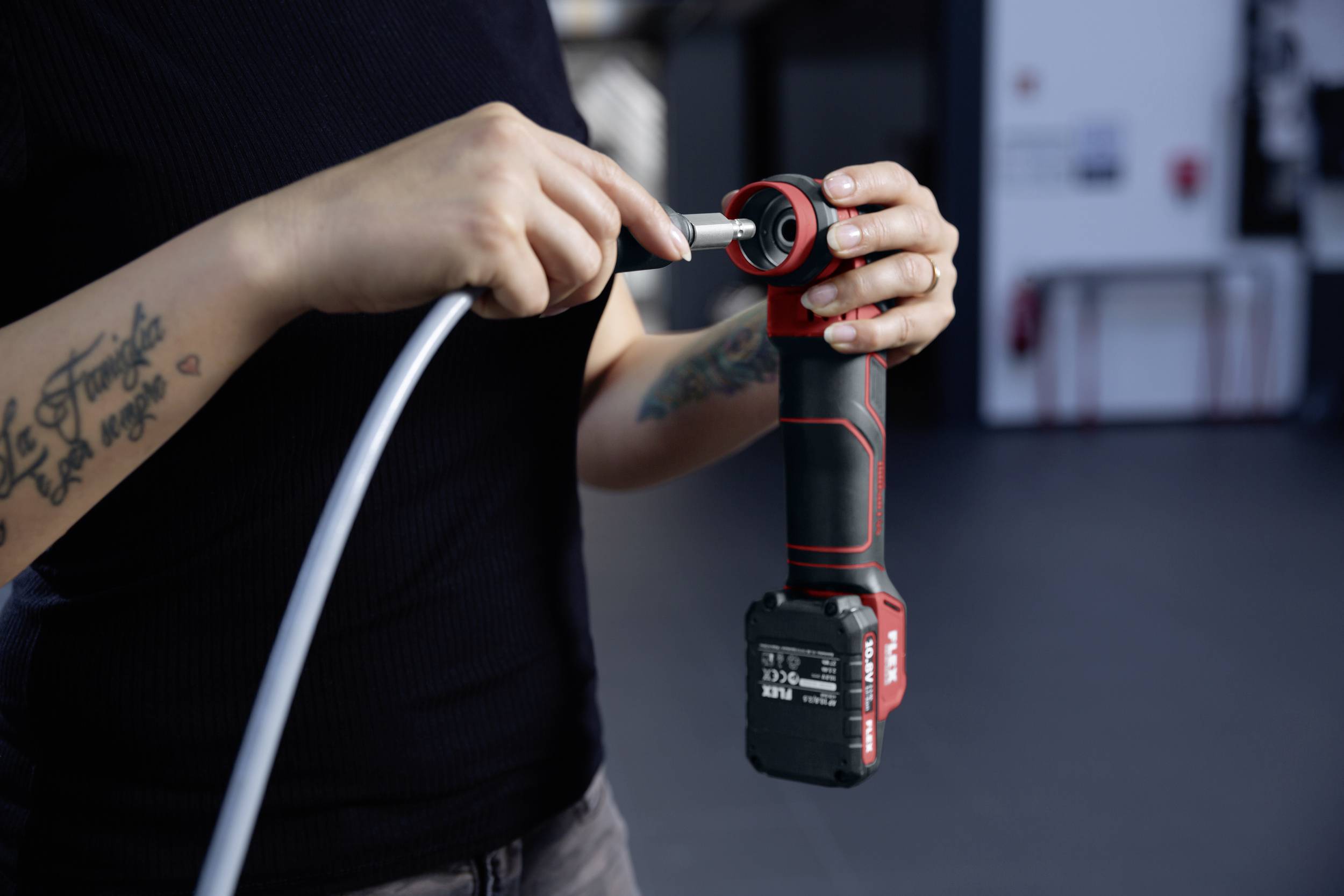 Jedna osoba drží červený, bezdrátový elektrický nástroj a připojuje k němu stříbrný kabel. Pozadí je rozmazané.
