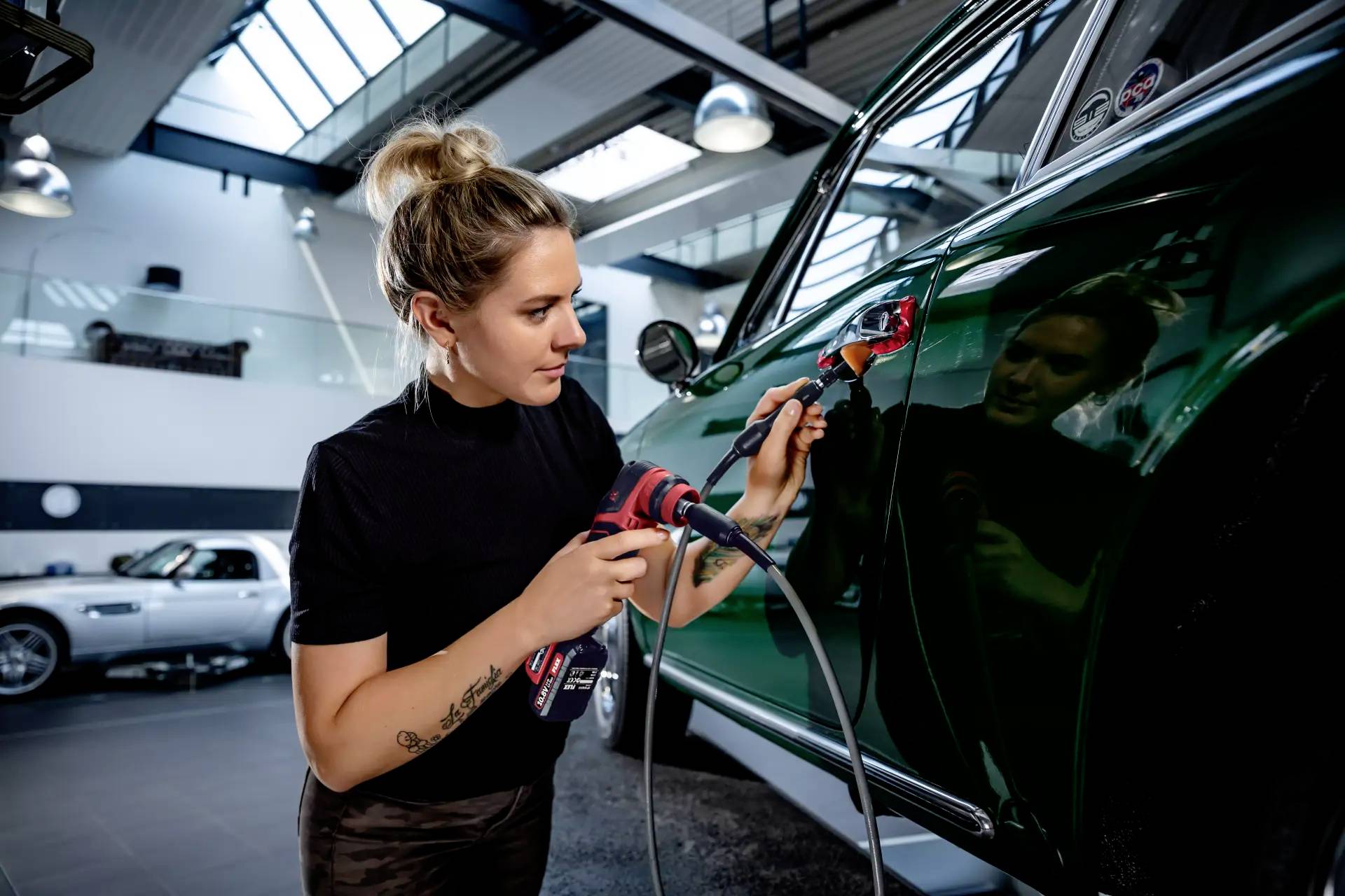 Žena leští automobil v moderní dílně. Používá leštičku na tmavě zelený lak. V pozadí jsou vidět další vozy.