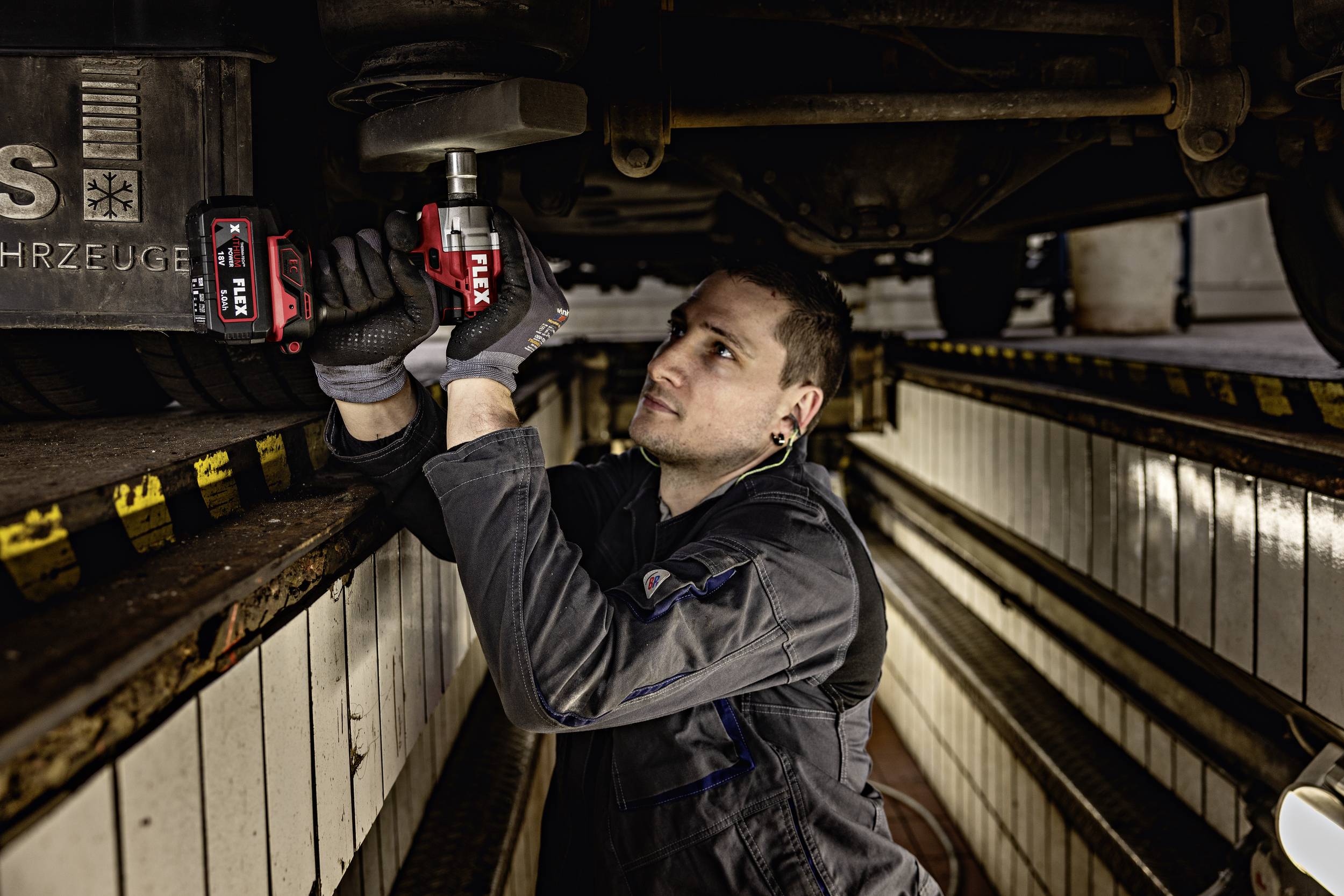 Mechanik pracuje pod vozidlem v montážní jámě. Používá elektrický nástroj k opravě některé z jeho součástí.