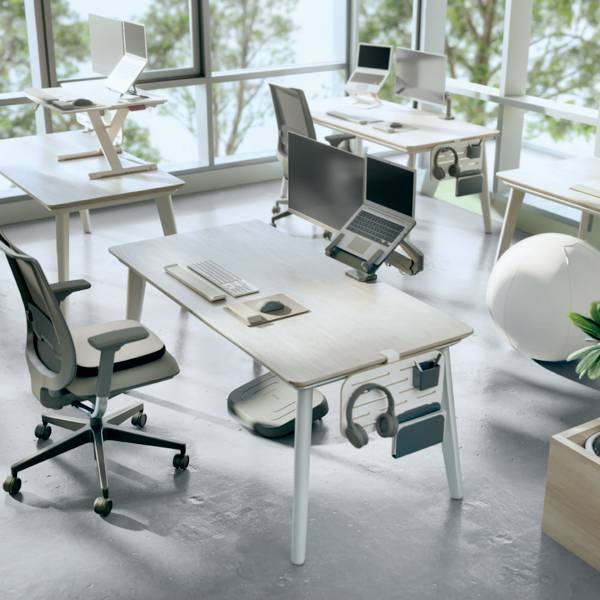 Několik moderních kancelářských stolů s laptopy, kancelářskými potřebami a ergonomickými židlemi v jasné, prostorné místnosti s velkými okny.