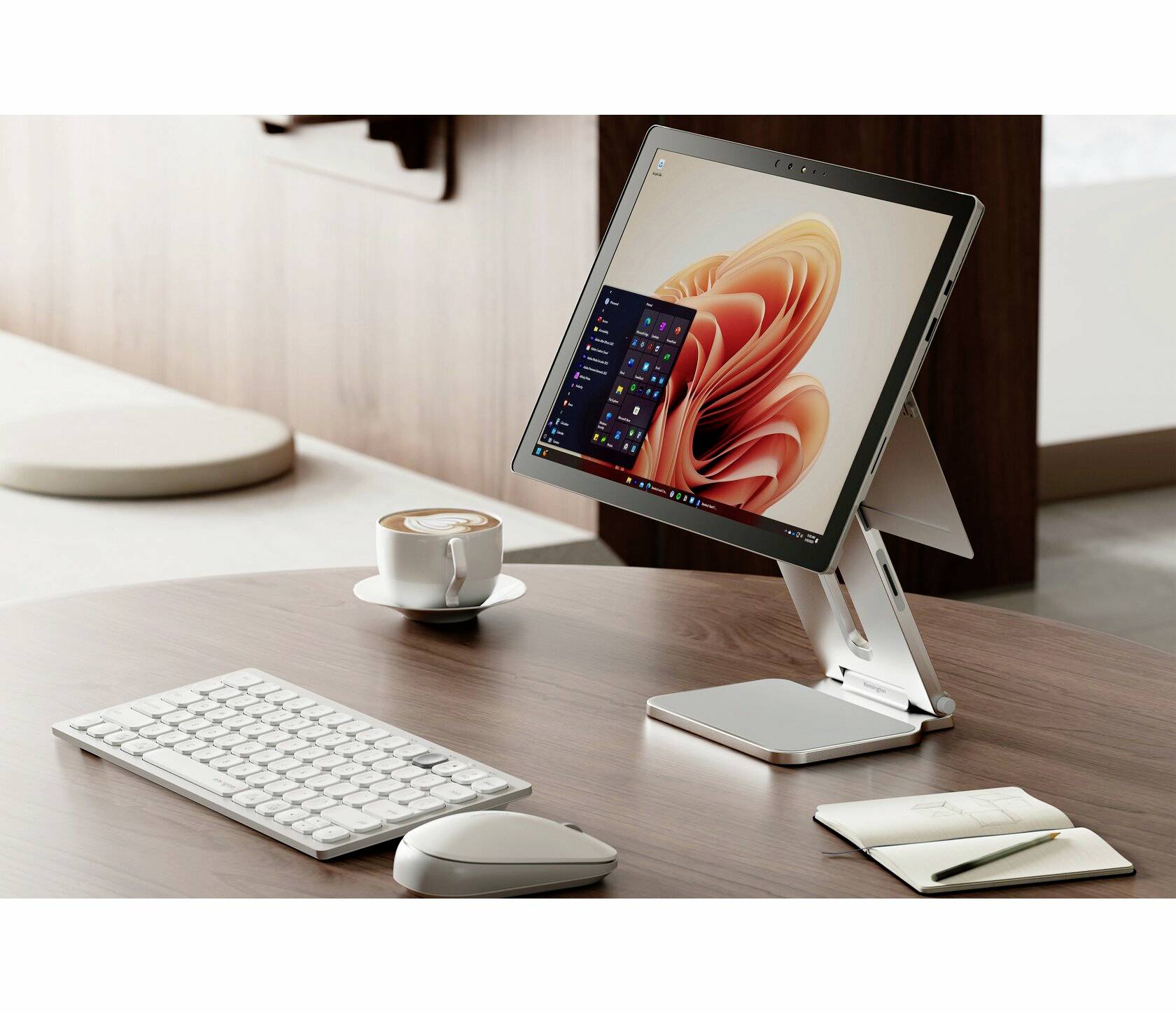 Tablet na stojanu zobrazuje operační systém s otevřenou spouštěcí lištou. Na stole jsou klávesnice, myš, poznámkový blok a šálek.