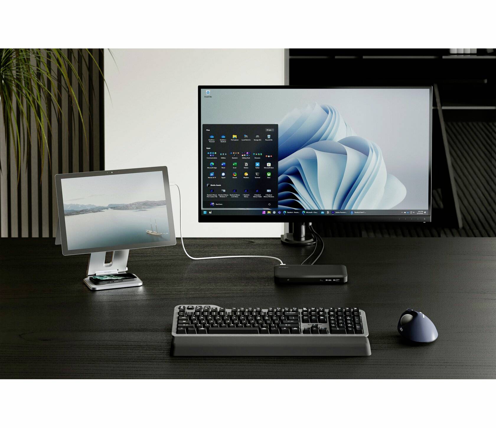 Černý psací stůl s černou klávesnicí, myší a dvěma monitory. Větší monitor zobrazuje plochu systému Windows.