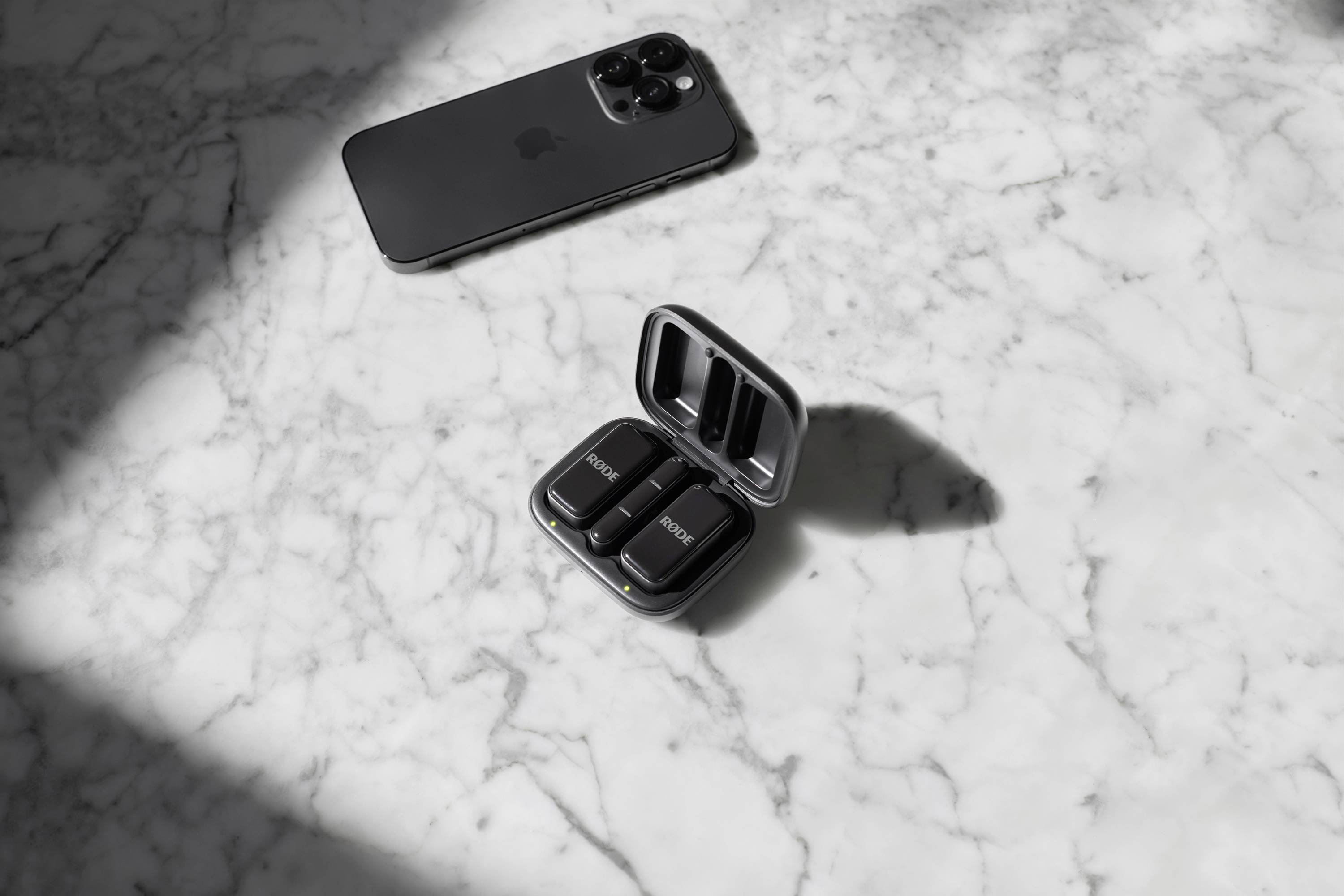 Černý smartphone a otevřený nabíjecí box se třemi kompaktními zařízeními leží na mramorové ploše v slunečním světle.