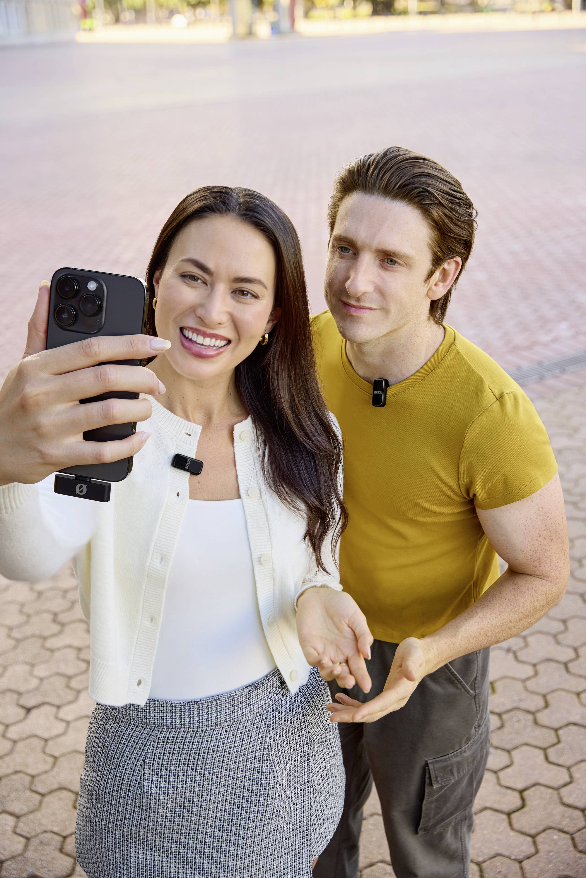 Žena a muž stojí venku a usmívají se do fotoaparátu smartphonu, který drží žena v ruce. Oba mají mikrofony.