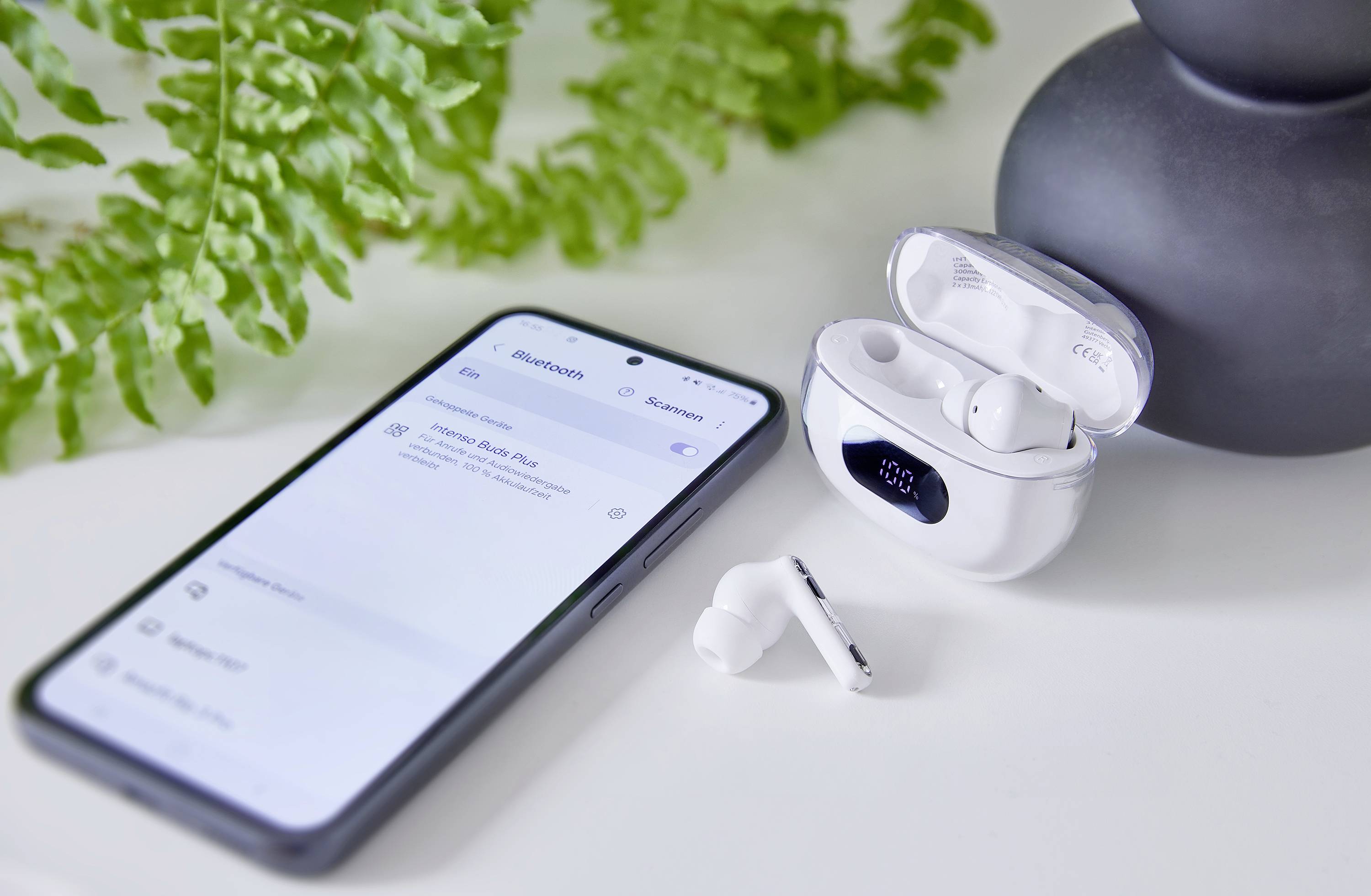 Smartphone vedle otevřeného pouzdra s bezdrátovými sluchátky, obklopené zelenými listy. Na displeji jsou zobrazeny nastavení Bluetooth.