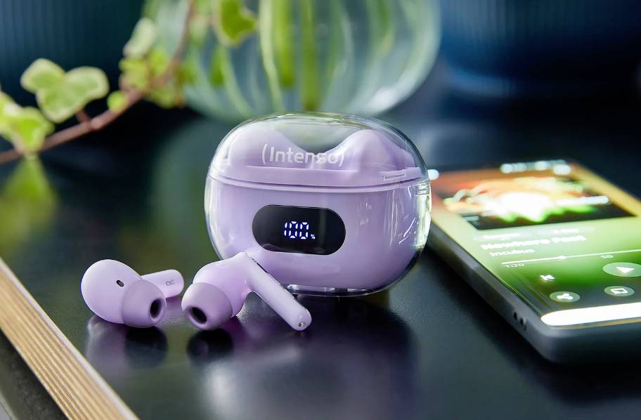 Fialové bezdrátové sluchátky v nabíjecím pouzdře leží na stole vedle smartphonu, který přehrává hudbu. Stav nabití sluchátek: 100 %.