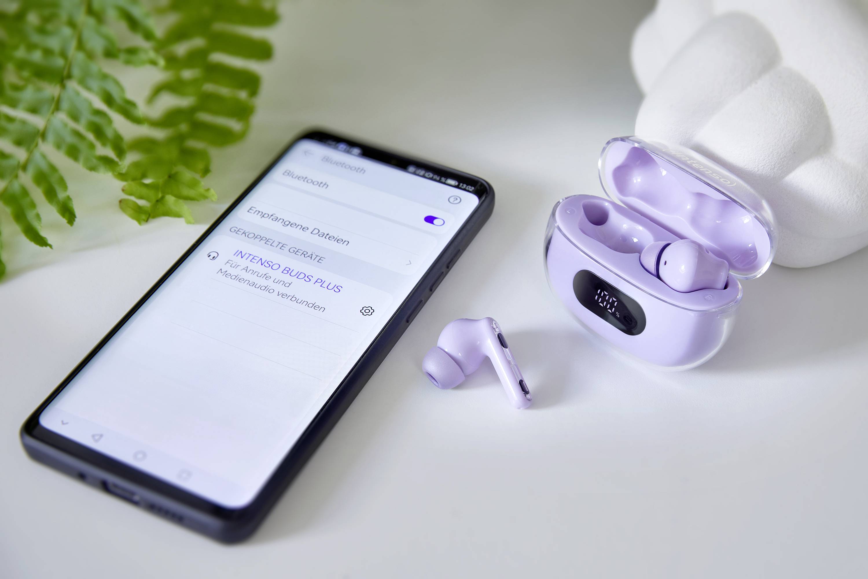 Smartphone vedle bezdrátových sluchátek v fialovém nabíjecím pouzdře. Na obrazovce nastavení Bluetooth s připojeným zařízením 'A78185 INDIE Buds'. Vlevo zelená rostlina v pozadí.