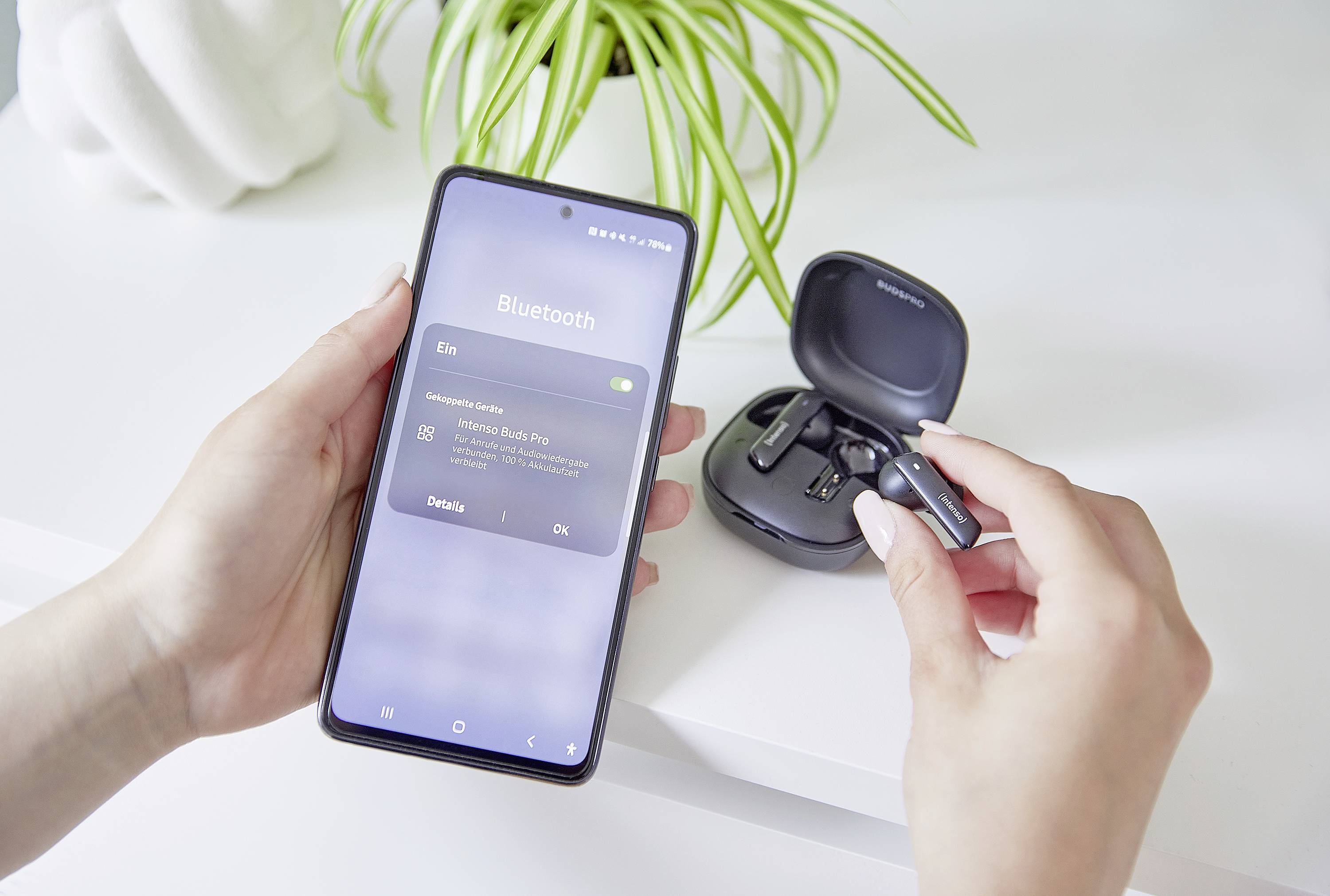 Jedna osoba připojuje bezdrátová sluchátka k mobilnímu telefonu pomocí Bluetooth. V pozadí je viditelná zelená rostlina.