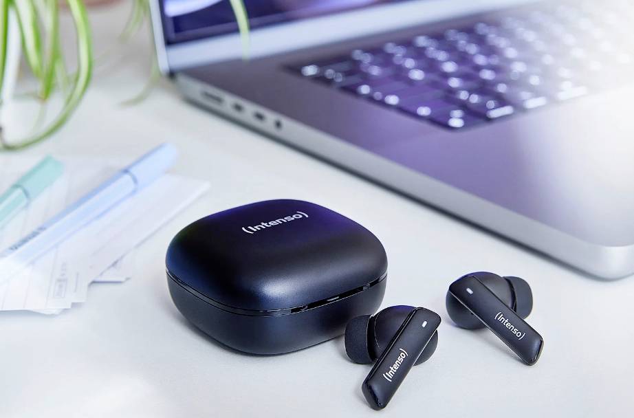 Bezdrátová černá sluchátka a pouzdro leží na bílém stole vedle otevřeného laptopku, v pozadí několik kancelářských potřeb.