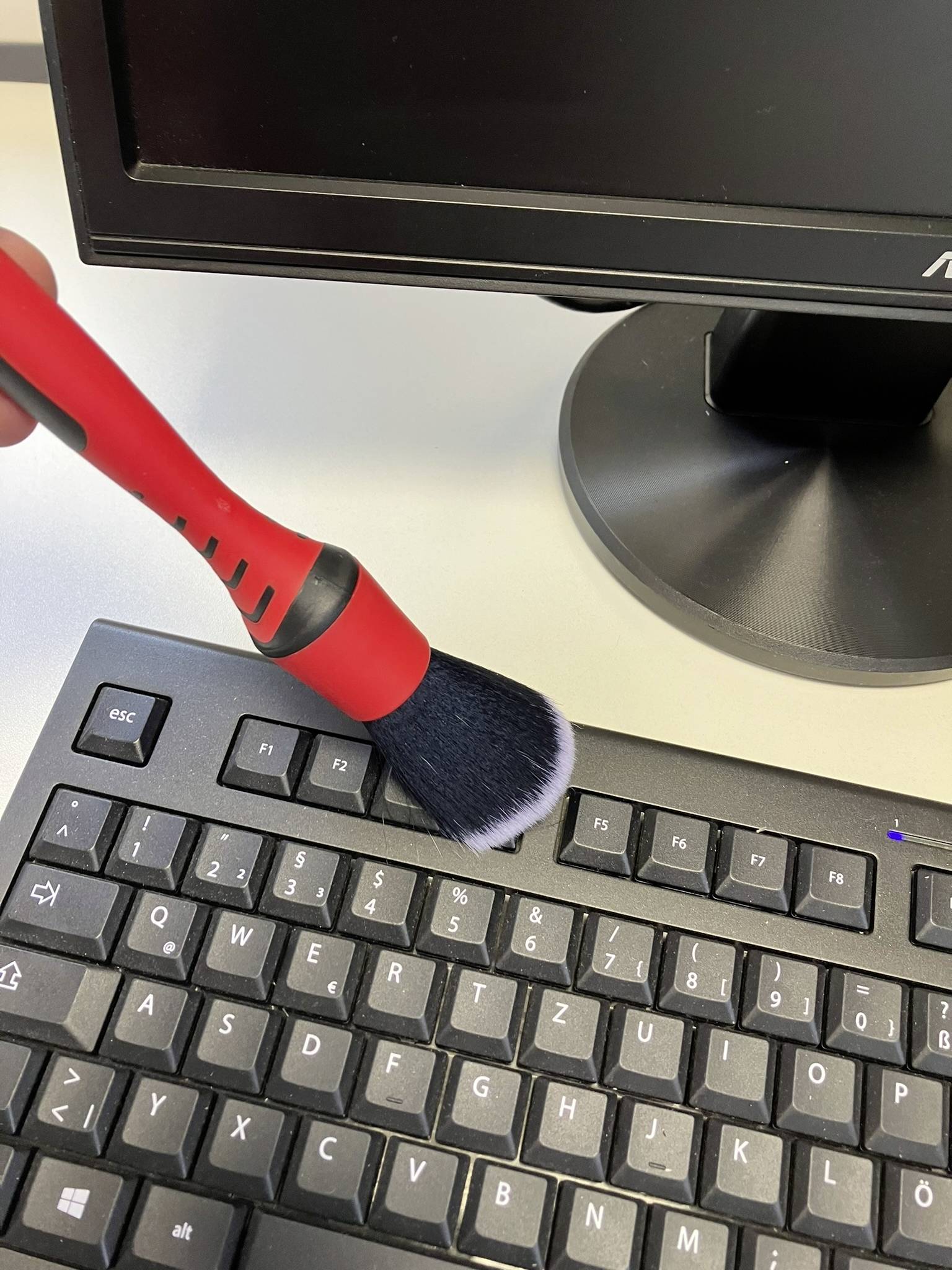 Ruka používá červeno-černý štětec k odstranění prachu z klávesnice počítače. V pozadí je vidět monitor.