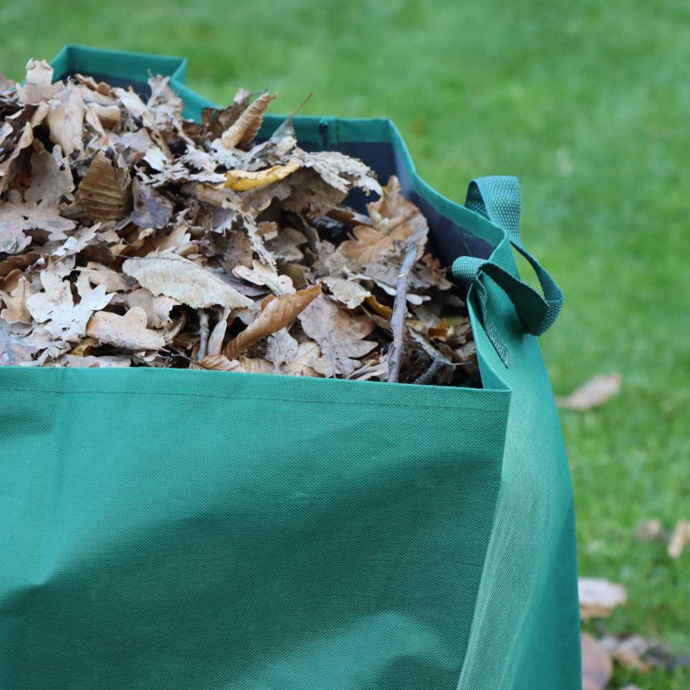Zelený pytel je naplněný až po okraj suchým podzimním listím; stojí na trávníku s roztroušenými listy v pozadí.