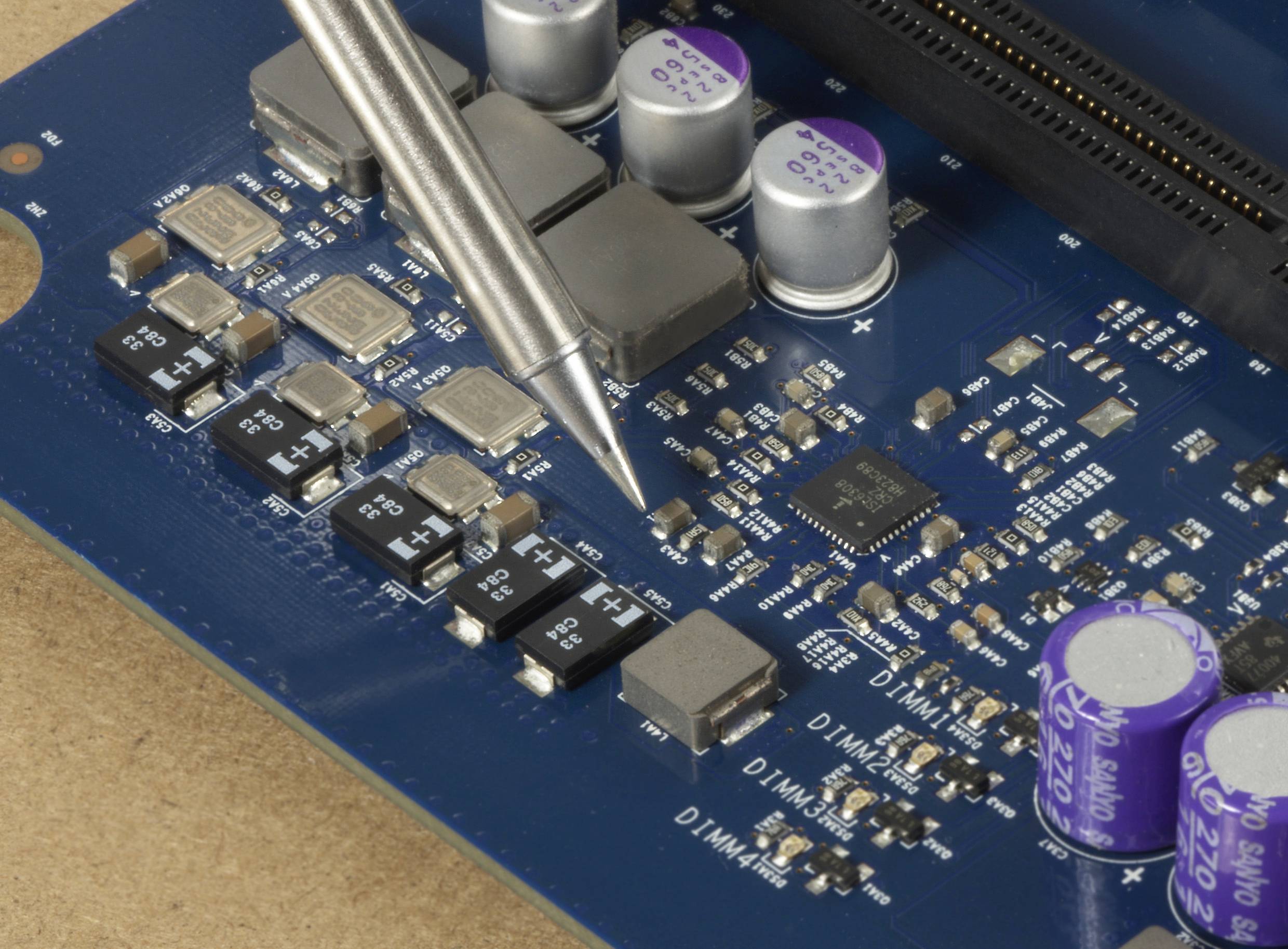 Detailní záběr modré desky plošných spojů s mikroči­pem a elektronickými komponenty. Pájecí pero se dotýká desky.