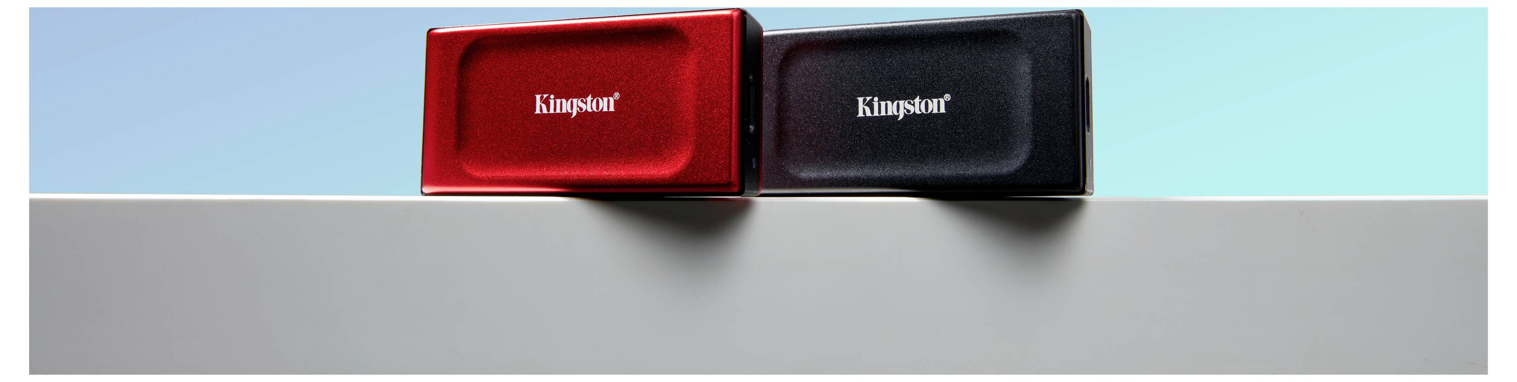 Dvě externí úložná zařízení v červené a černé barvě s nápisem 'Kingston' leží vedle sebe na šedém povrchu před modrým pozadím.