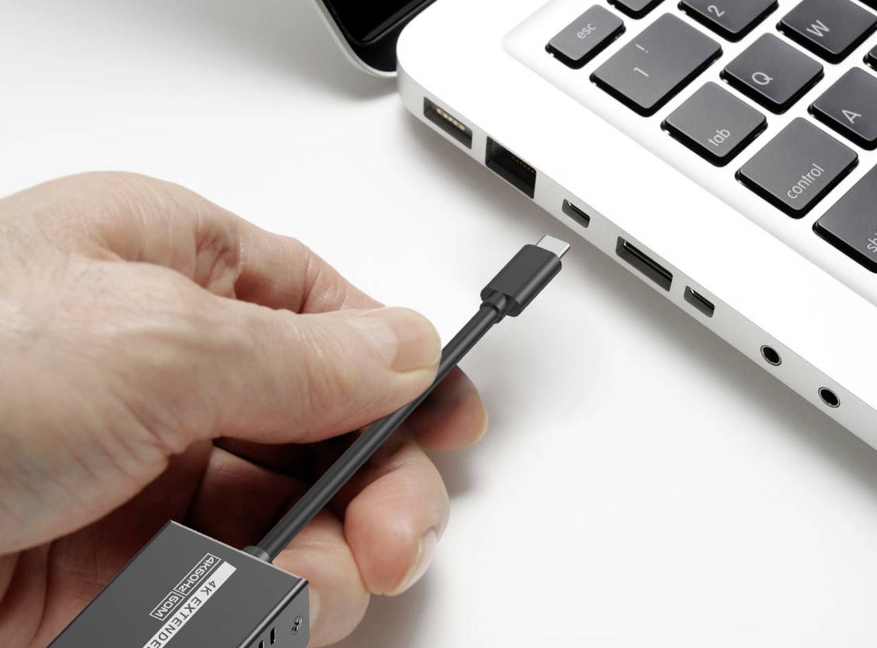 Ruka zasunuje USB-C kabel do laptopu, který leží na stole. Laptop má po straně několik portů.