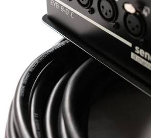 Černé audio zařízení s několika XLR konektory a kabely; slouží k propojení mikrofonů nebo audiovybavení.