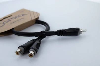 Černobílý cinch Y-kabel se dvěma zdířkami a jednou zástrčkou, ležící na bílém povrchu, propojený s papírovou krabičkou.