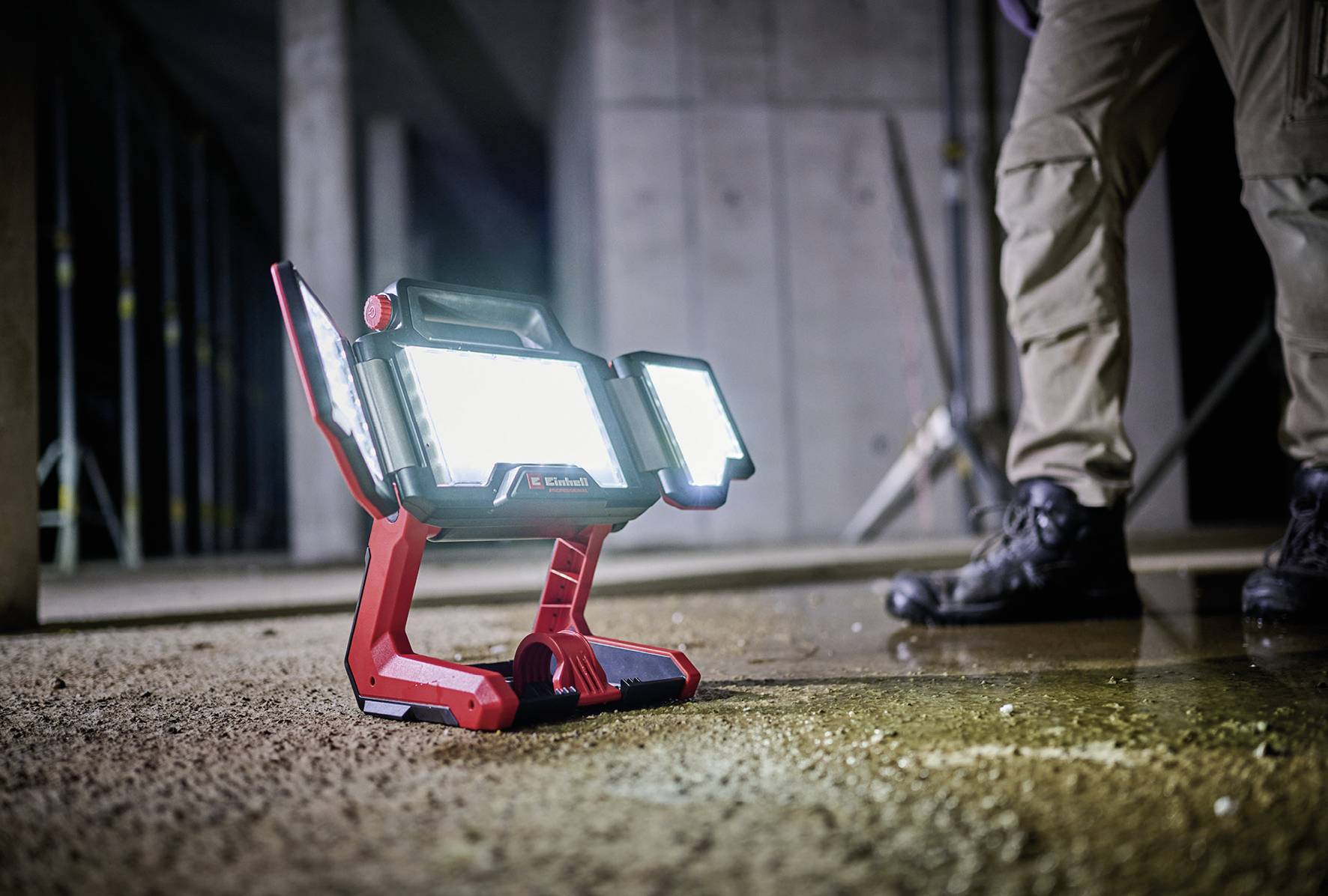 Jasná stavební lampa svítí v polotmavé místnosti. V pozadí jsou vidět nohy osoby v pracovním oblečení.