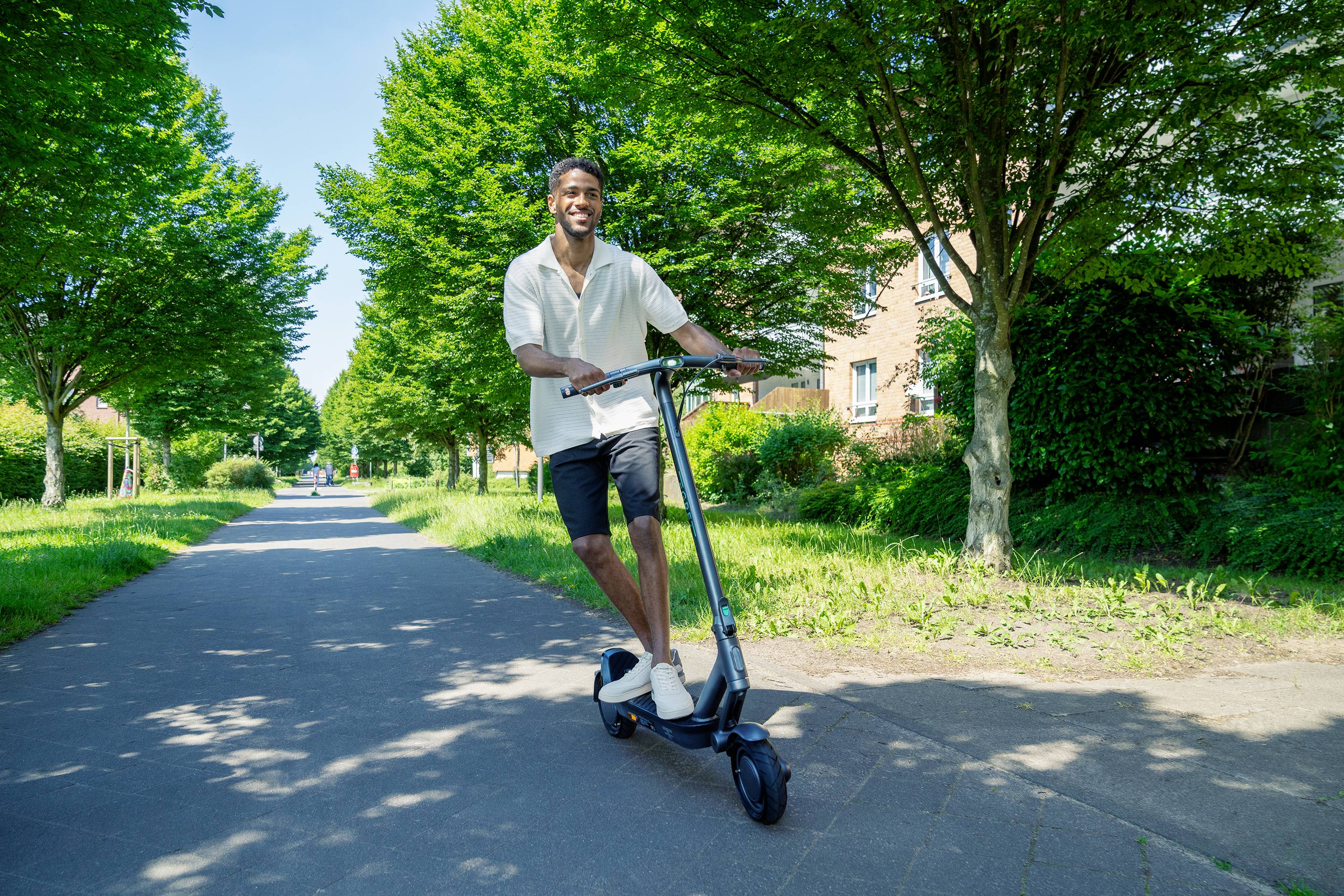 Muž vesele jede na elektrickém koloběžce podél cesty lemované stromy. Svítí slunce a obloha je modrá.