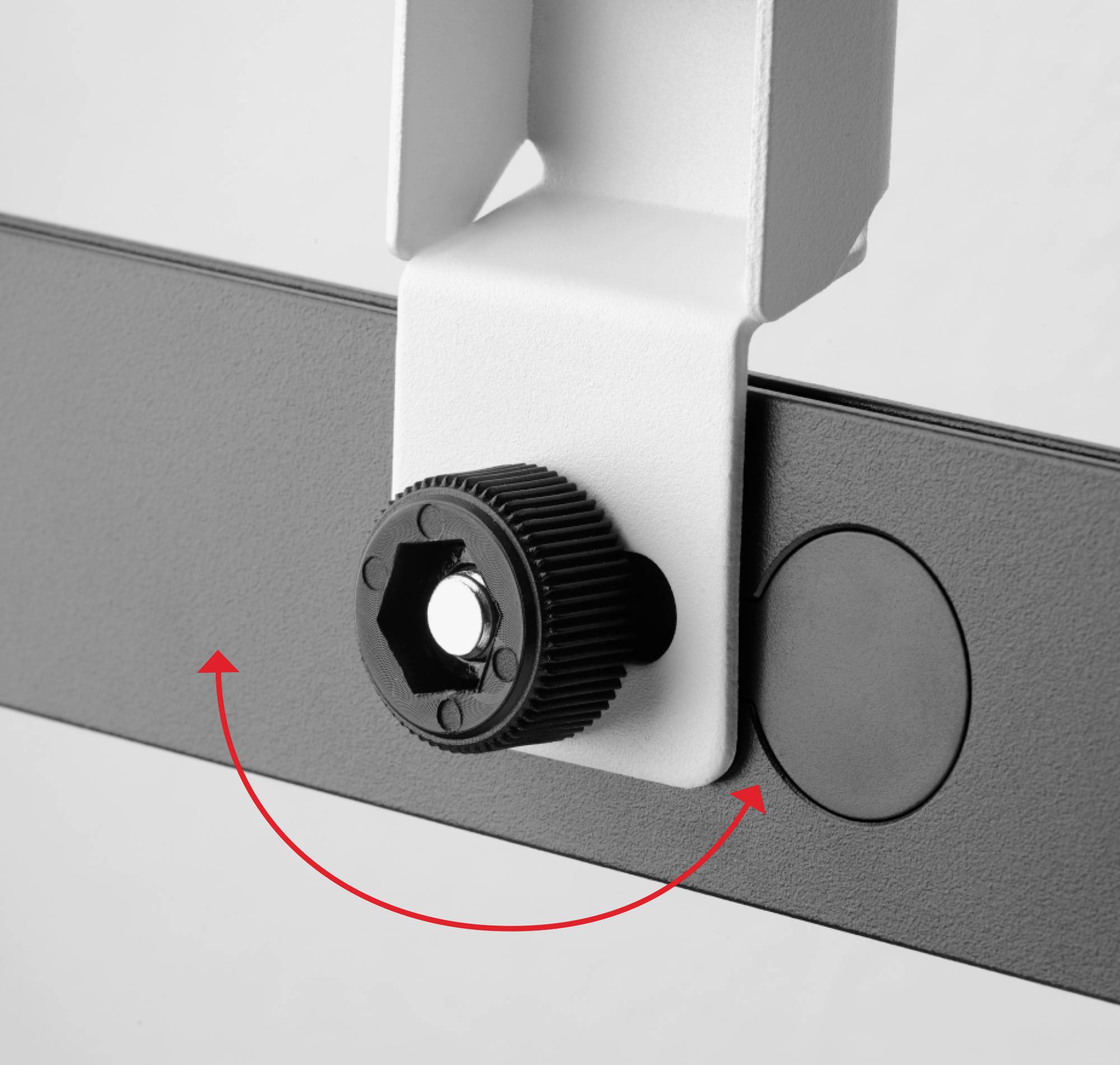 Detailní záběr černého šroubu upevněného v bílém držáku. Červený šipka ukazuje směr otáčení šroubu.
