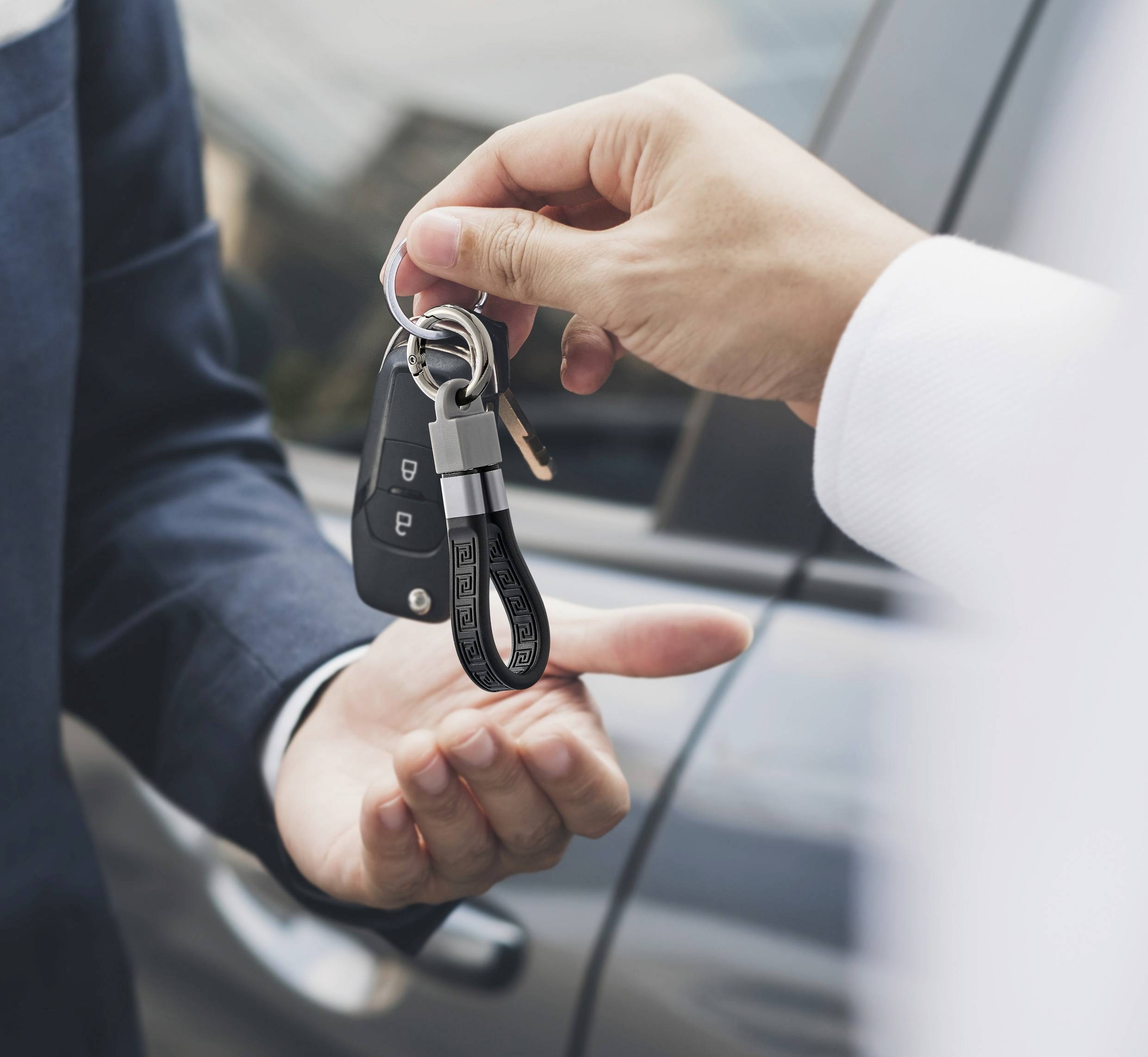 Jedna osoba předává druhé klíč od auta, v pozadí je vidět automobil.
