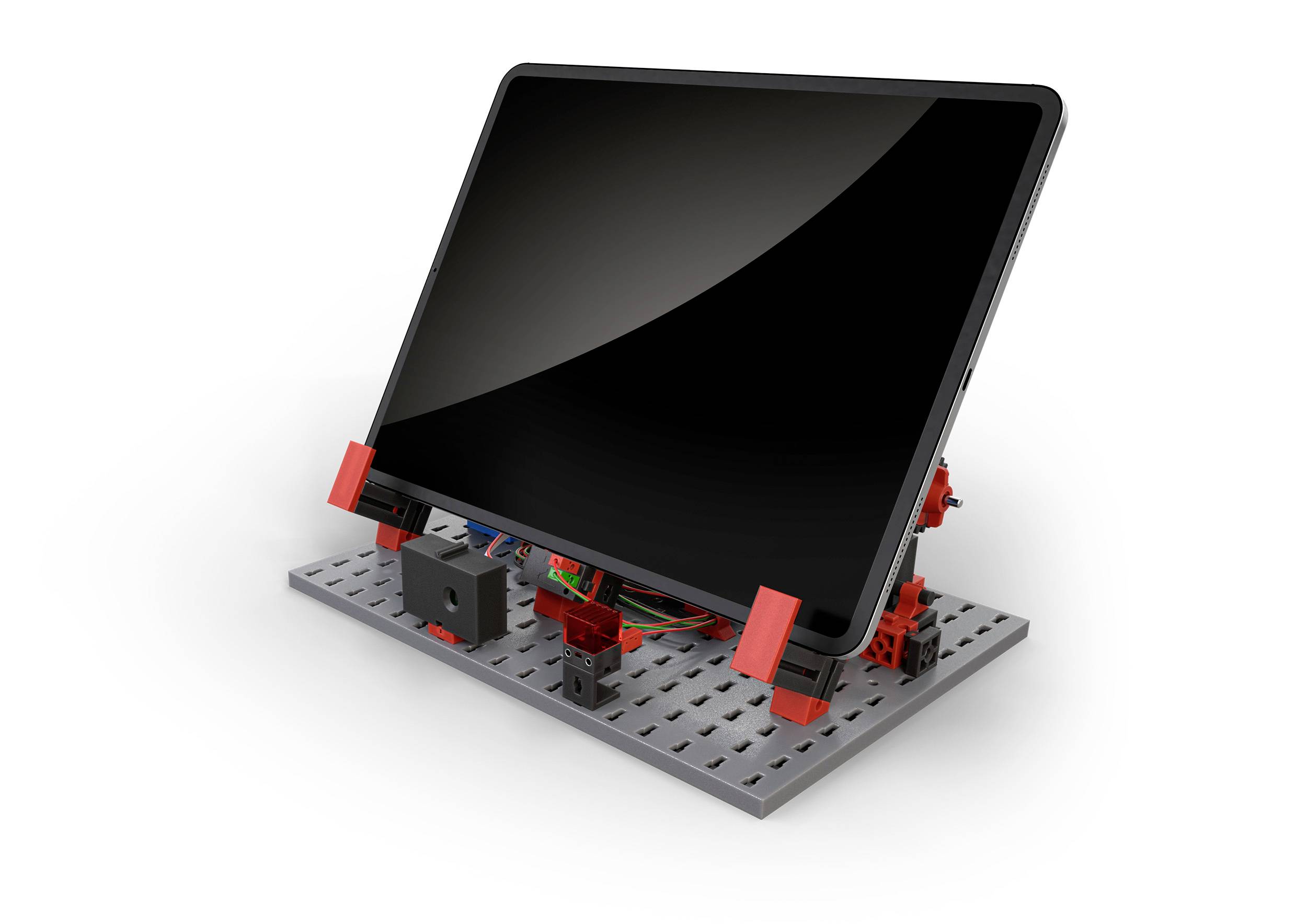 Tablet na naklápěcím stojanu, připevněný na šedé perforované desce s červenými a černými úchyty.