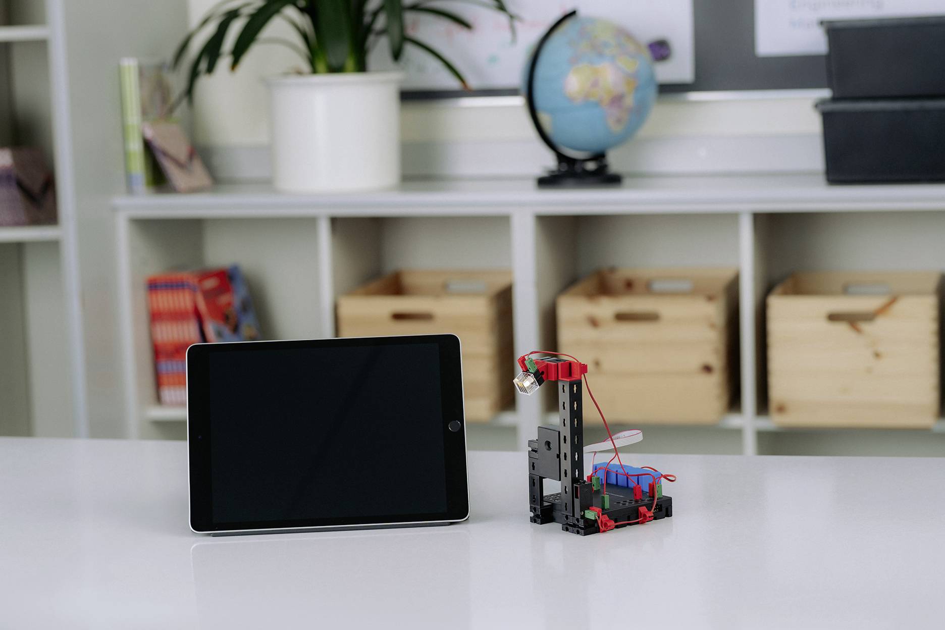 Tablet a malý robot na stole ve třídě. V pozadí police s rostlinami a glóbus.