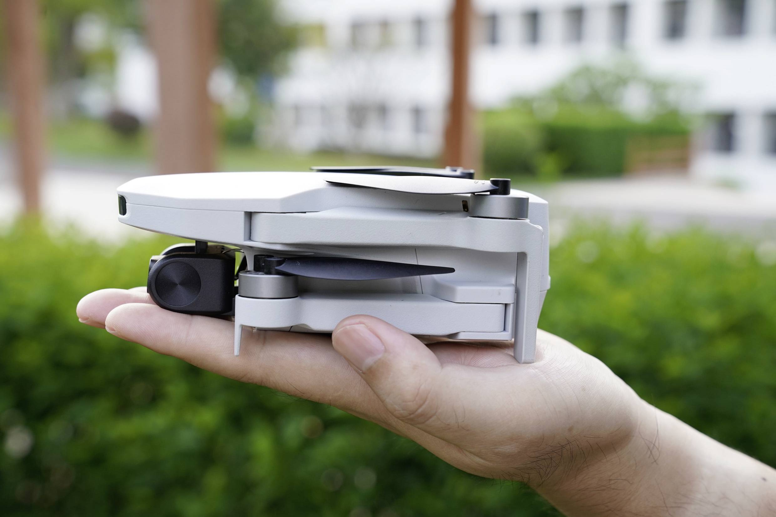 Jedna ruka drží malou složenou dron venku. V pozadí jsou rozmazaně viditelné stromy a budovy.