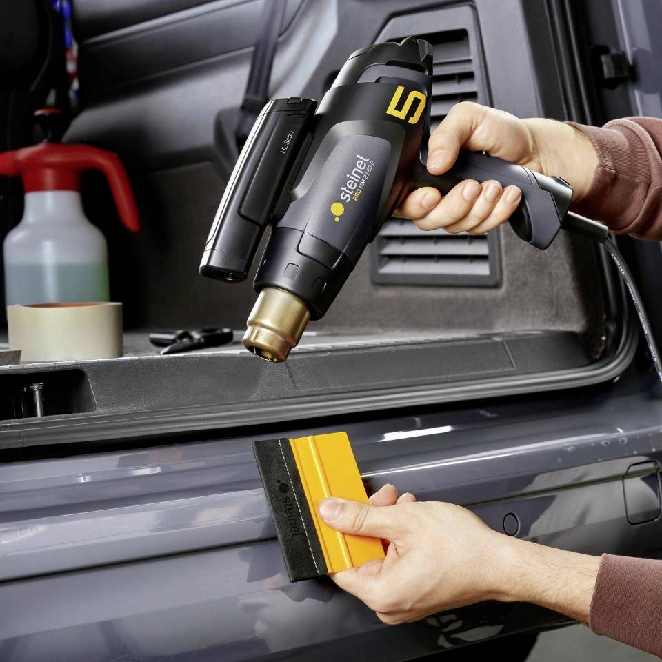 Osoba drží černý horkovzdušný ofukovač a žlutou stěrku u zadní části automobilu. V pozadí jsou viditelné nástroje.
